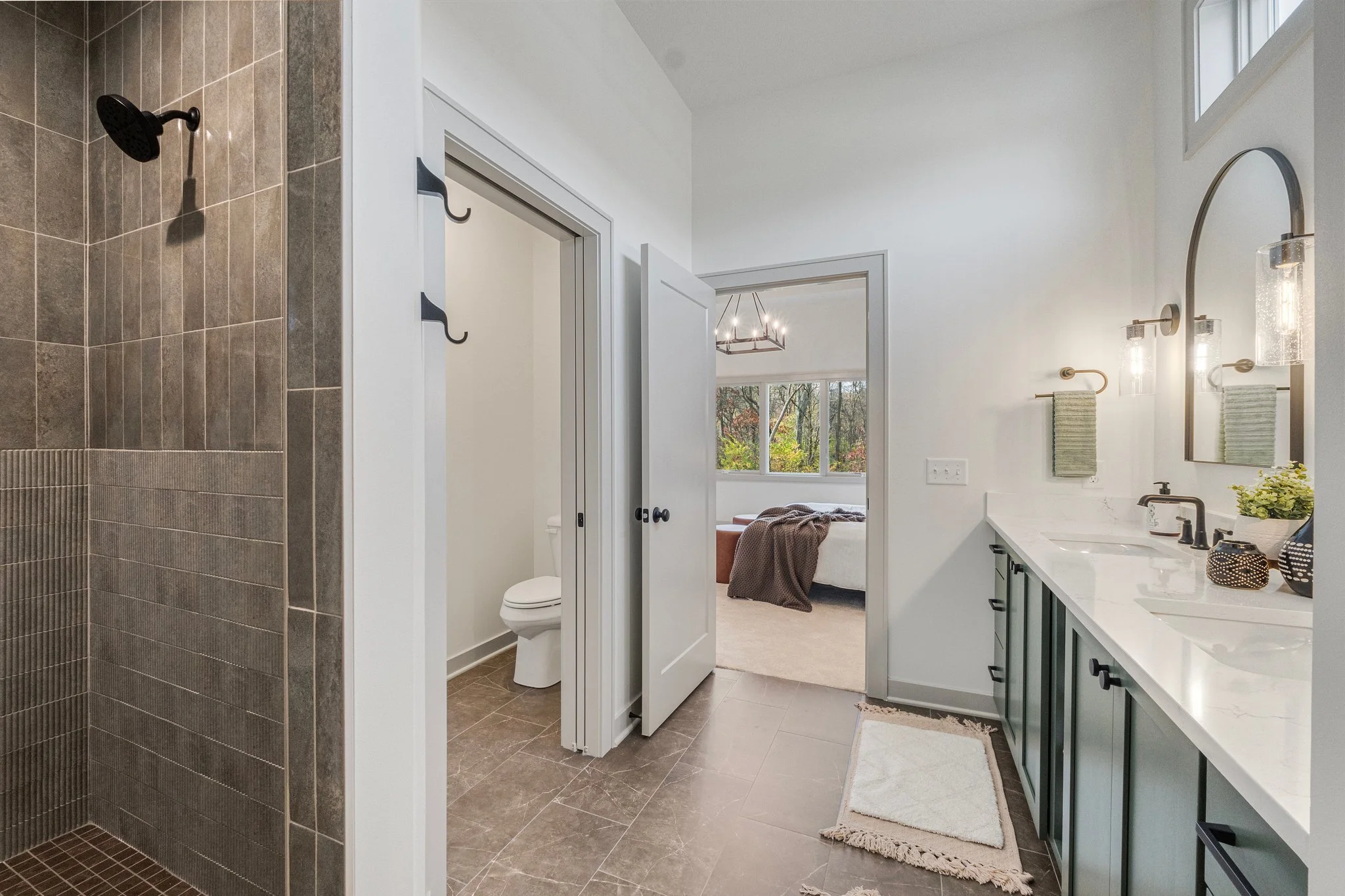 Modern bathroom with a tiled shower, a toilet area separated with a door, a double vanity with green cabinet doors, mirrored medicine cabinets, and window with a view of a bedroom with a bed, large window, and chandelier.