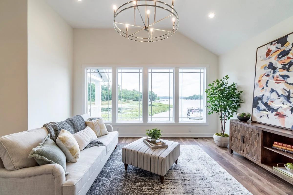 Living room with a large window showing a lakeside view, a beige sofa with pillows, a striped ottoman, a wooden console with a colorful abstract painting, a potted plant, and a modern circular chandelier.