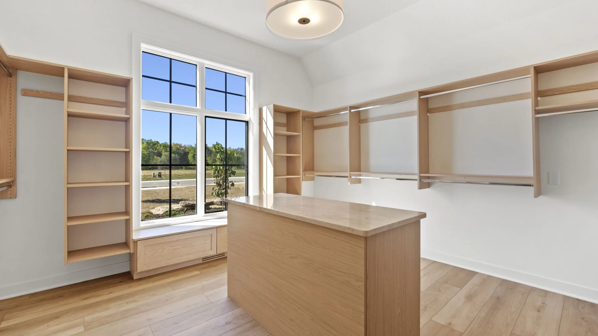 Empty walk-in closet or dressing room with built-in wooden shelves, a central wooden island with a marble top, and large window with a view of outdoors.