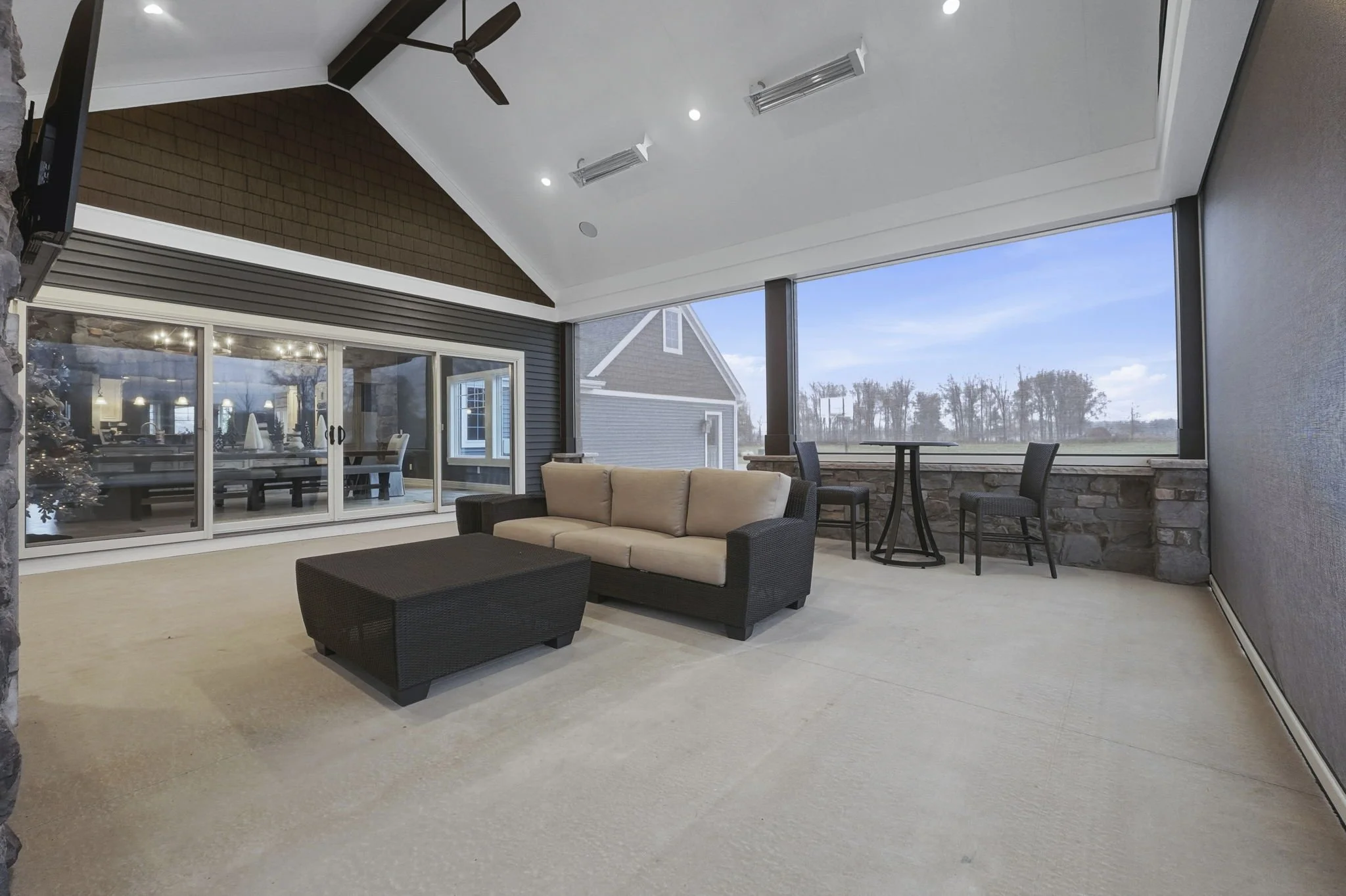 A spacious indoor patio with beige carpet, black wicker sofa with beige cushions, small coffee table, and two chairs with a small round table in front of a large glass sliding door. Outside view of a grassy field and trees, with a stone wall along th