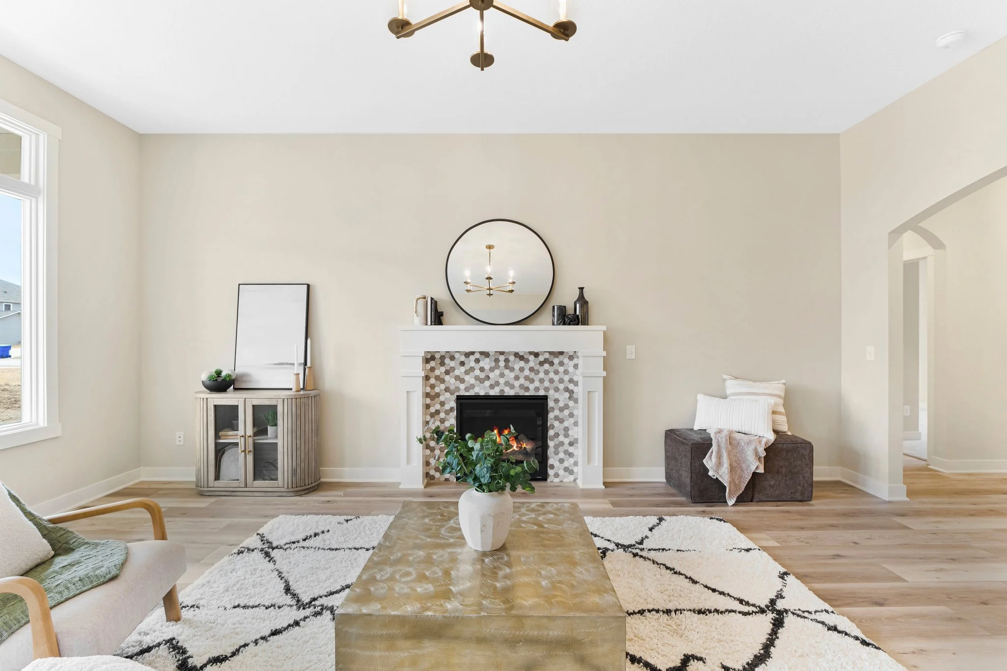 Living room with a beige wall, a white fireplace with decorative tiles, a round mirror above it, a plant on a gold coffee table, and a black ottoman with pillows to the right. There is a side cabinet with decor on the left and a window on the left wa