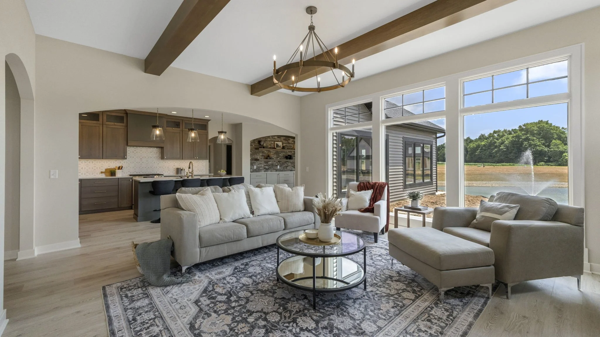 Living room with large windows overlooking a pond and fountain, beige sofa, armchair, round coffee table, hardwood flooring, and an open view of the kitchen with modern cabinetry and pendant lights.