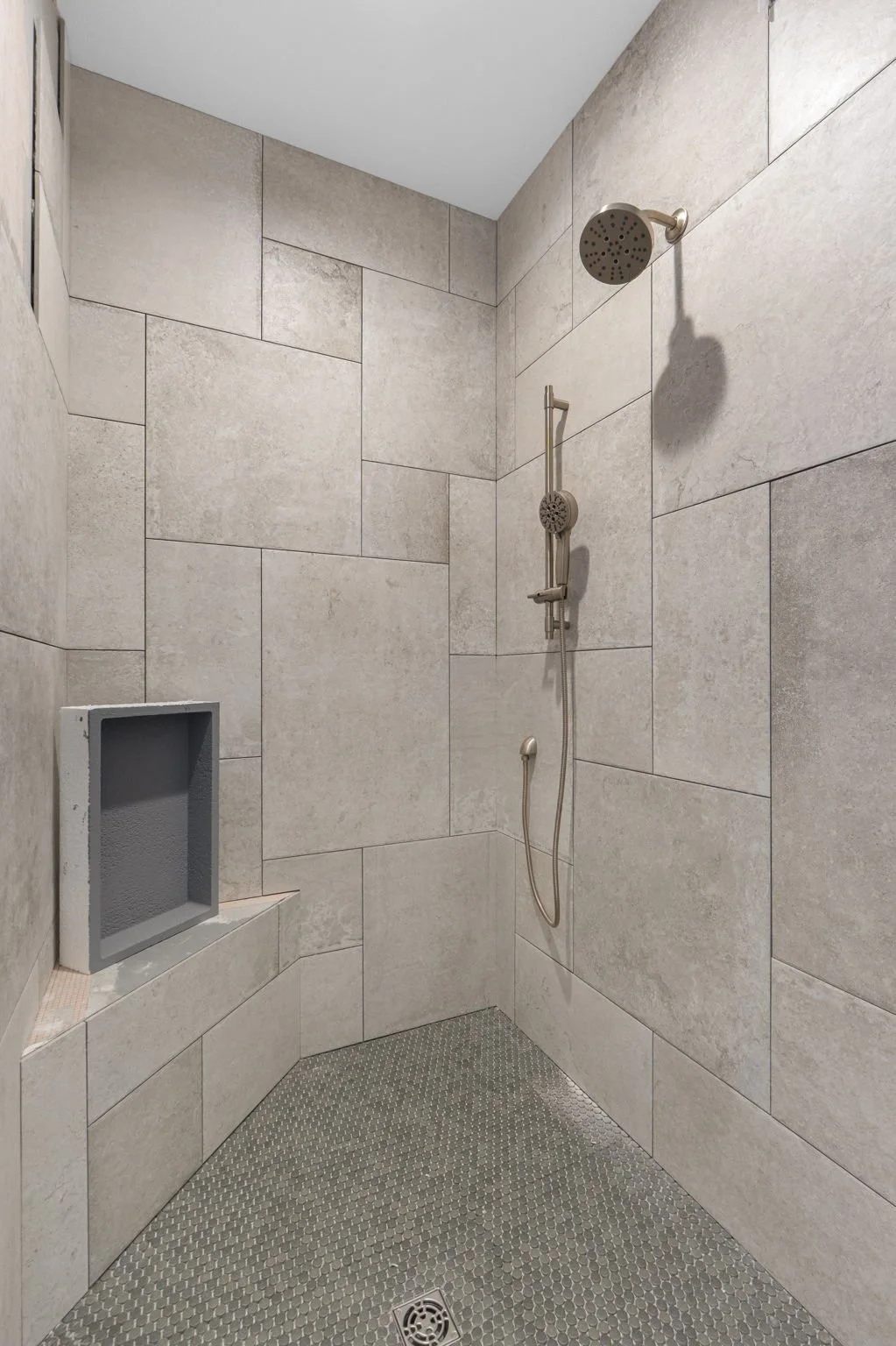 A modern shower with large beige tiles on the walls, a silver showerhead, a hand-held shower wand, a built-in niche, and a small drain on the floor.