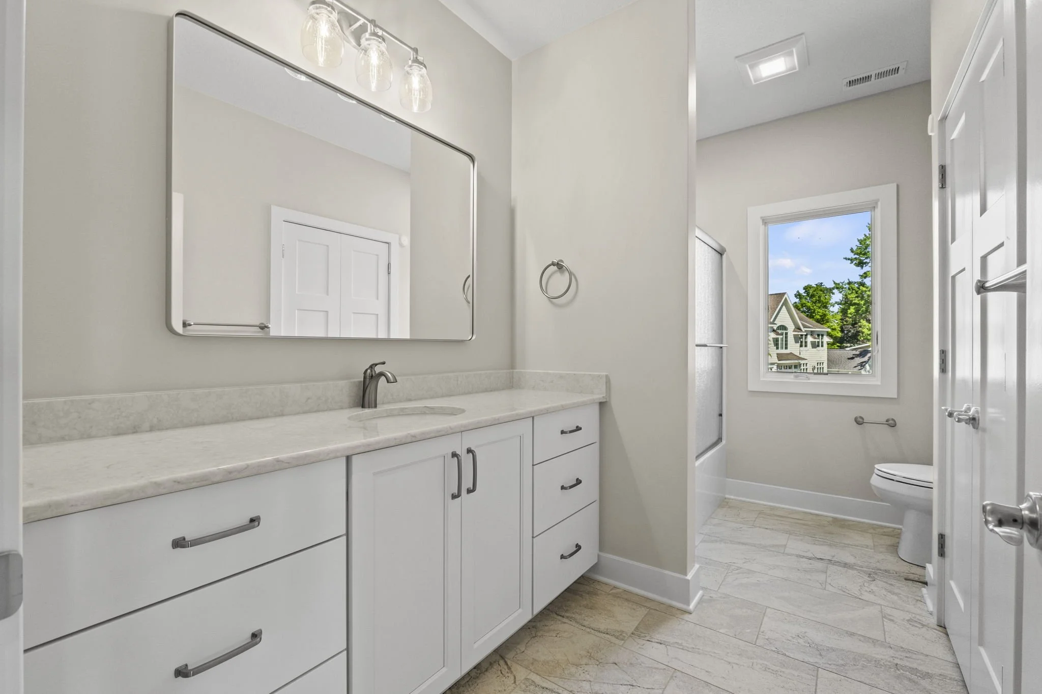 Bathroom with white cabinetry, marble countertop, large mirror, and a window showing houses and trees outside.