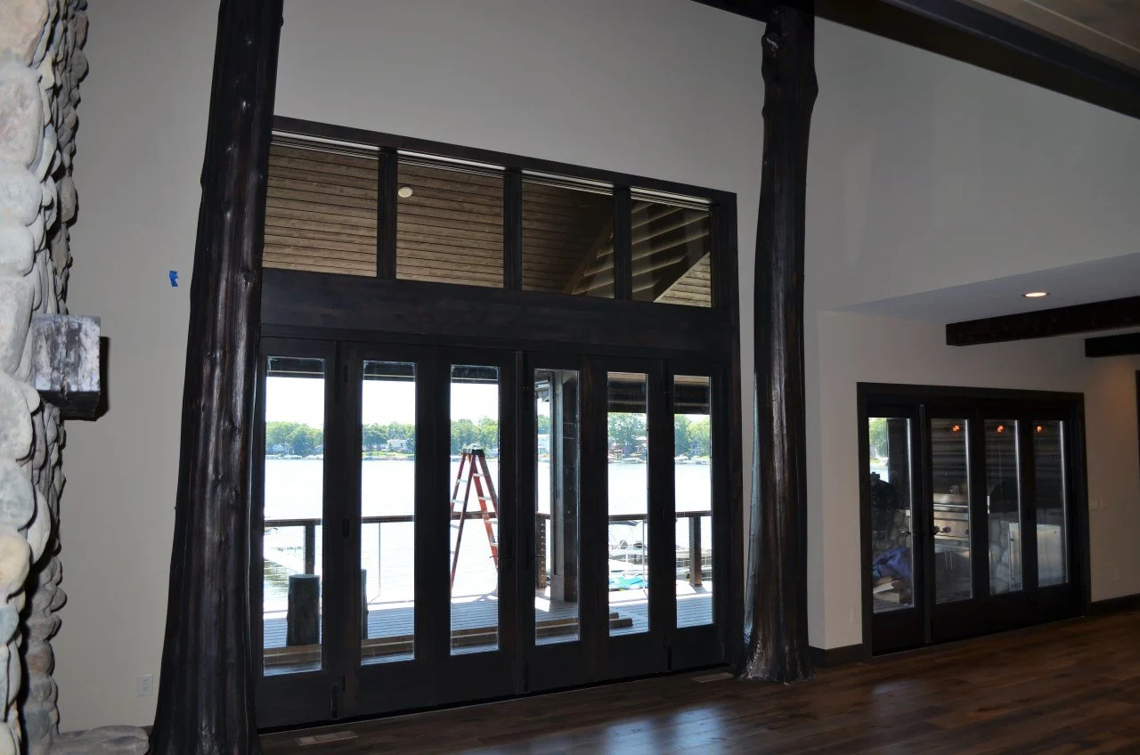 Interior view of a room with large black-framed glass doors leading to an outdoor deck, overlooking a body of water. The room features a stone wall on the left and dark wooden flooring. There is a ladder visible outside on the deck.