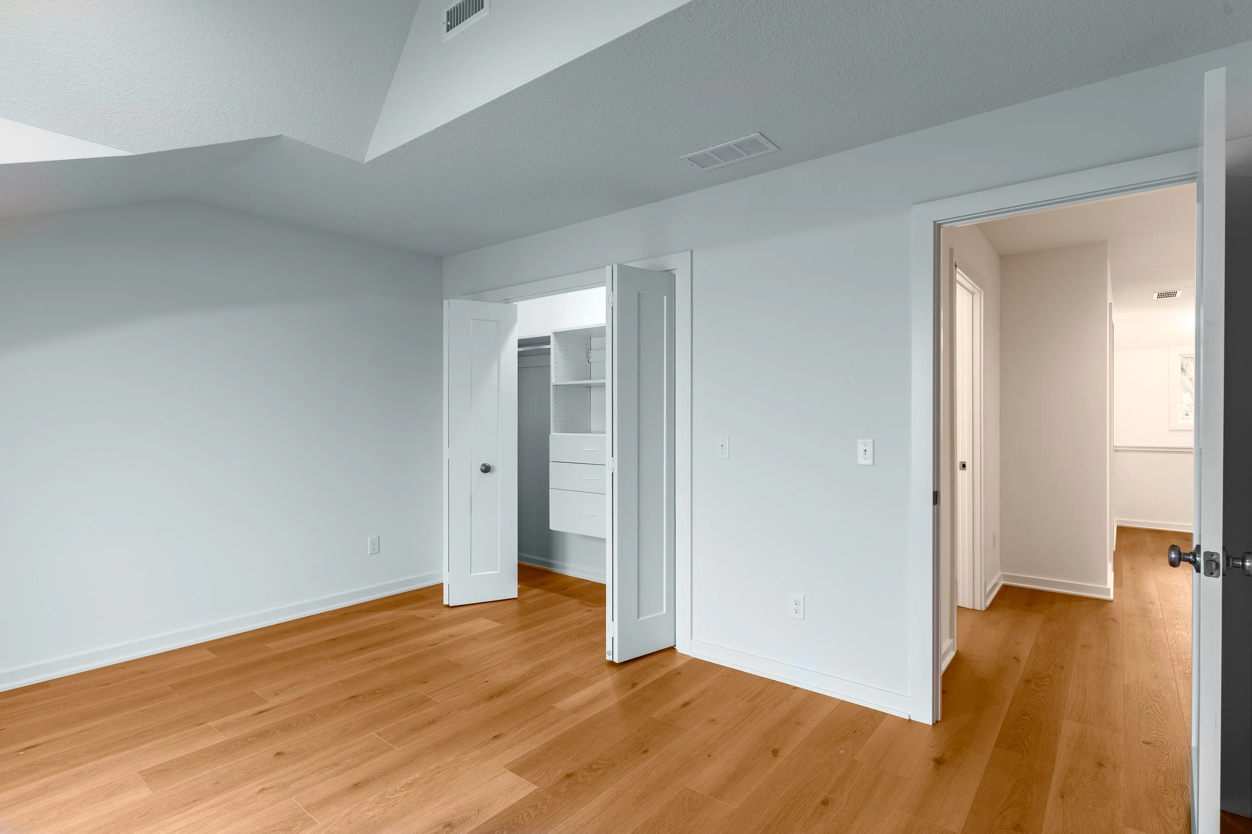 Empty room with white walls and wooden floors, featuring a closet with open doors and an adjoining hallway.