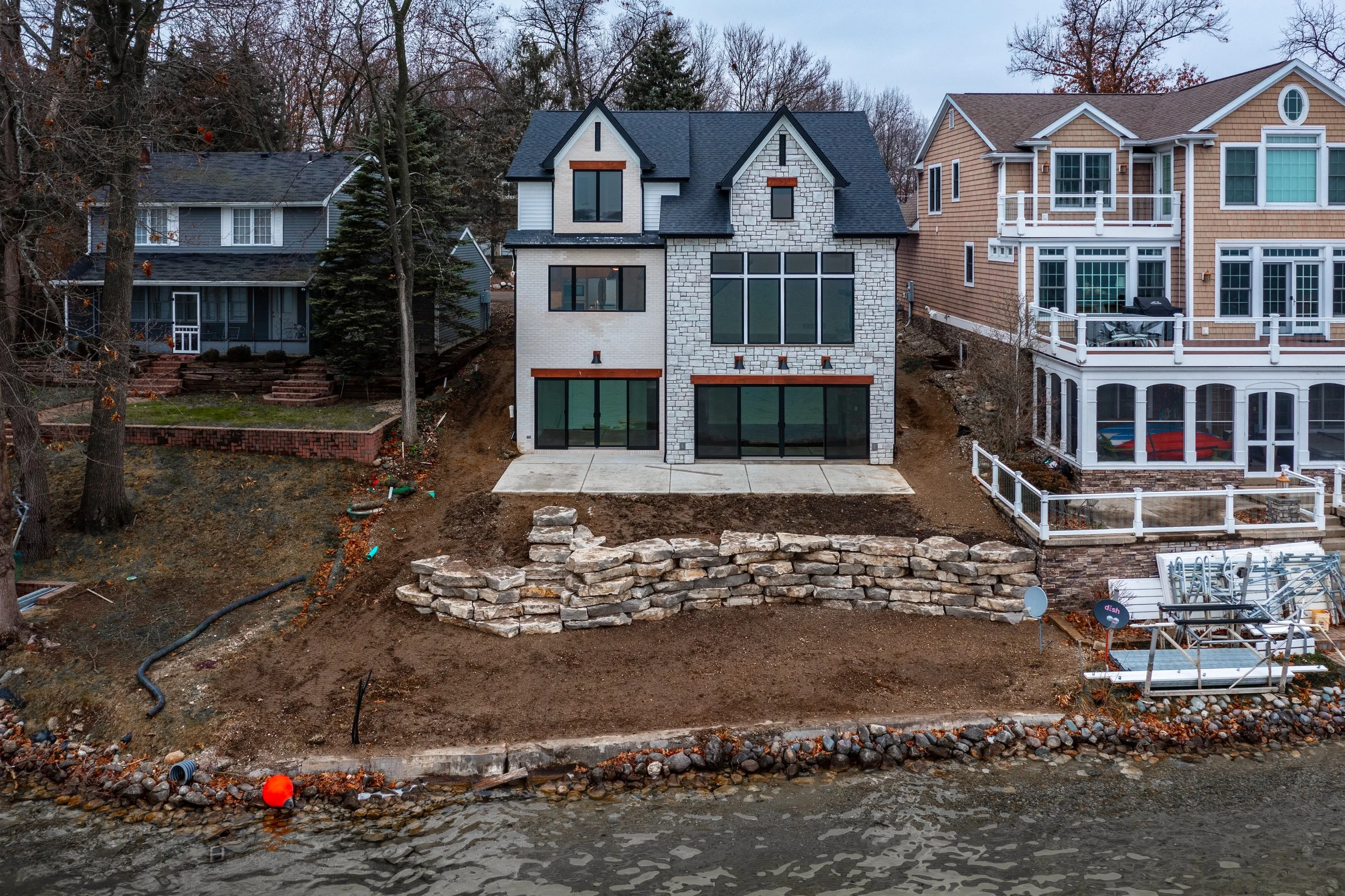 Newly constructed multi-story house along a waterfront with a stone retaining wall, large glass windows, and a concrete patio, surrounded by neighboring houses and trees.