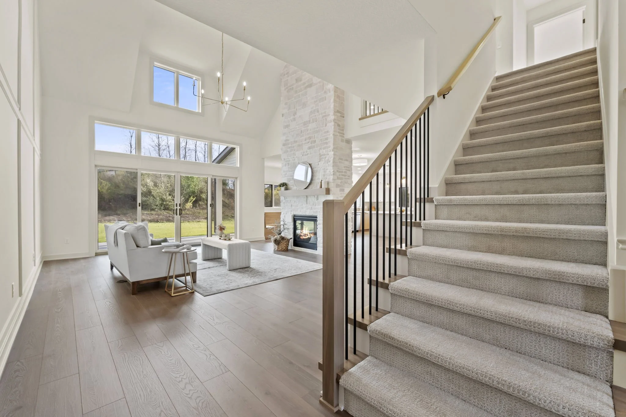 Bright living room with large windows, white walls, a fireplace, a white sofa, and a staircase with light carpet leading to the upper floor.