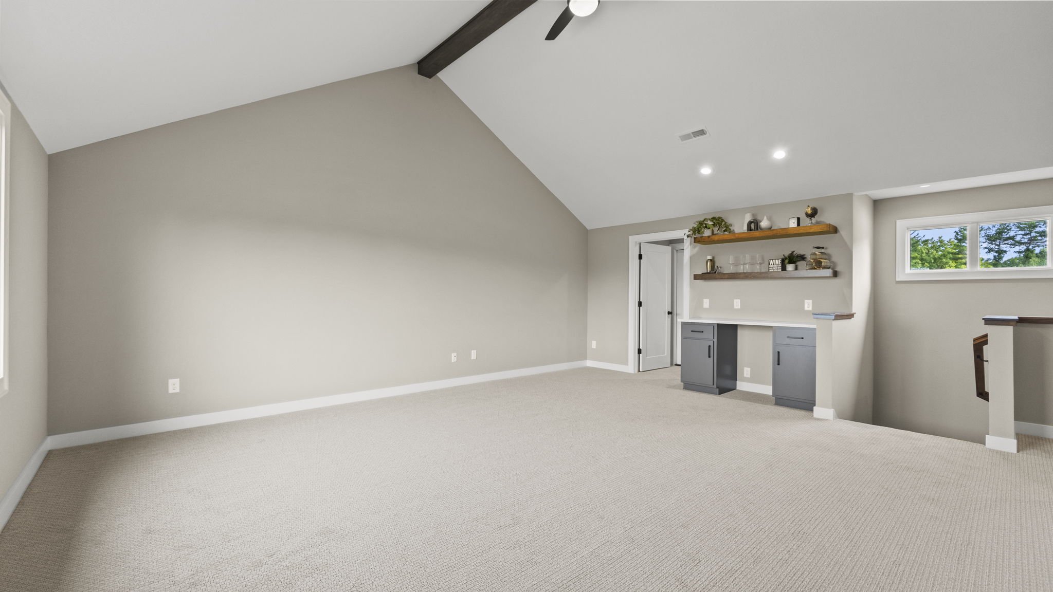 Empty living room with beige carpet, light gray walls, a small built-in desk with gray cabinets, two floating shelves with decorations, a window showing trees outside, and a ceiling fan with a black beam.