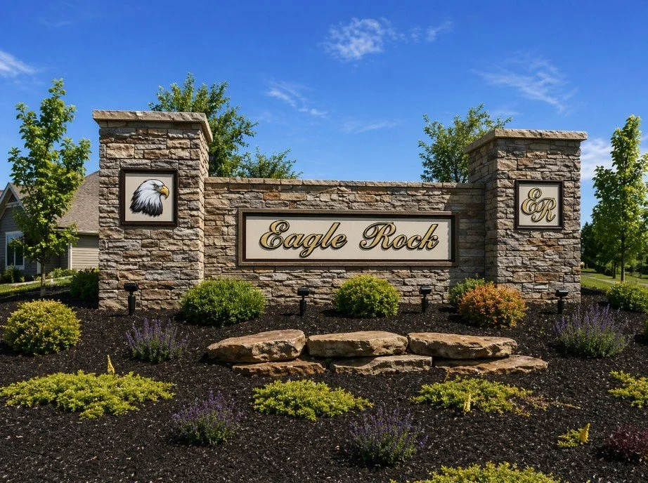 A stone entrance sign for Eagle Rock, with a painted eagle head logo on a small side pillar, and the initials 'ER' on another side pillar. The sign is surrounded by landscaped bushes and flowers under a bright blue sky.