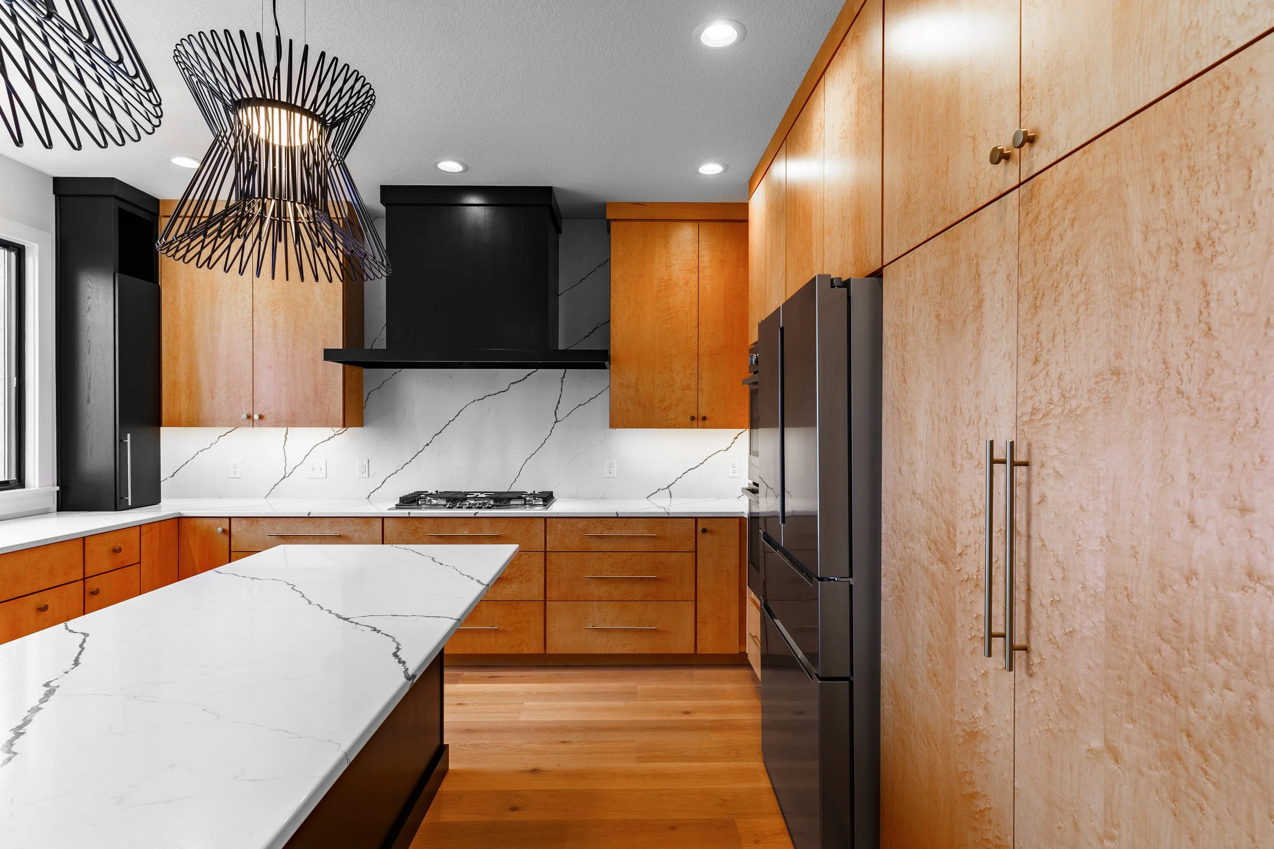 Modern kitchen with wooden cabinets, white marble countertops, black kitchen hood, stainless steel refrigerator, and black wire pendant lighting fixture.