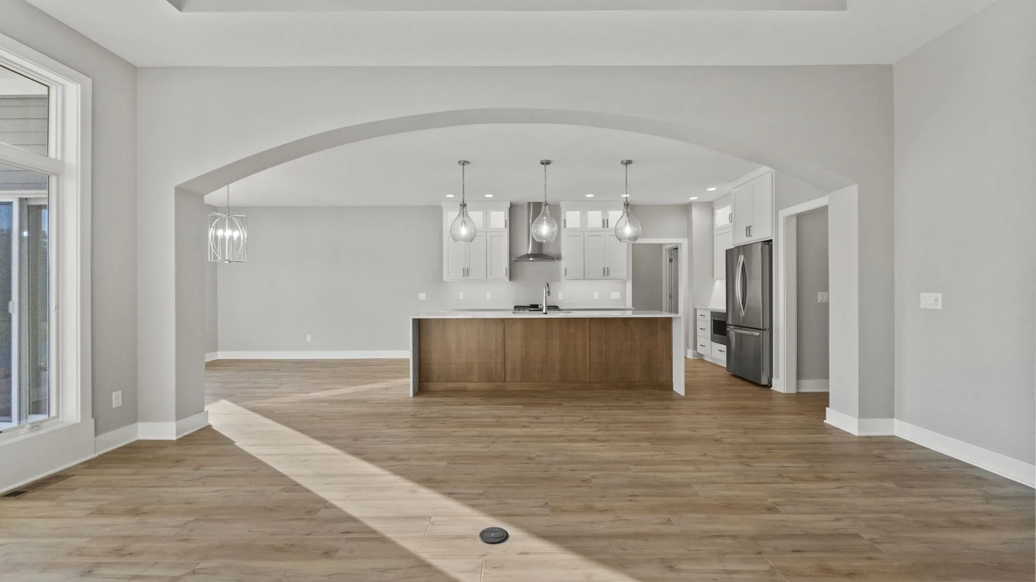 Empty modern kitchen with wooden flooring, white cabinets, stainless steel appliances, large island with wood finish, three pendant lights, and bright natural light coming from a sliding glass door.