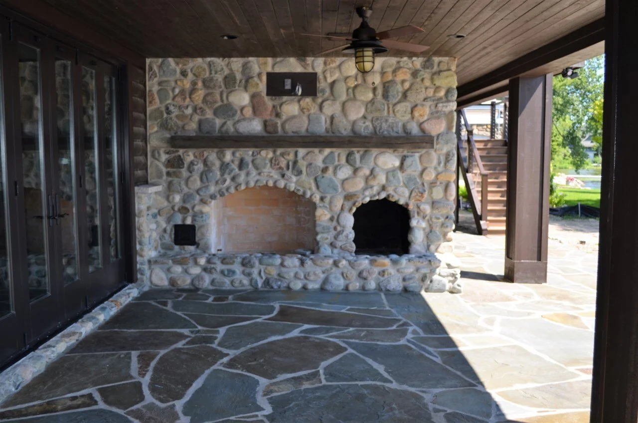 Outdoor patio area with a stone built fireplace, stone flooring, a ceiling fan, and wooden stairs leading to an upper level.
