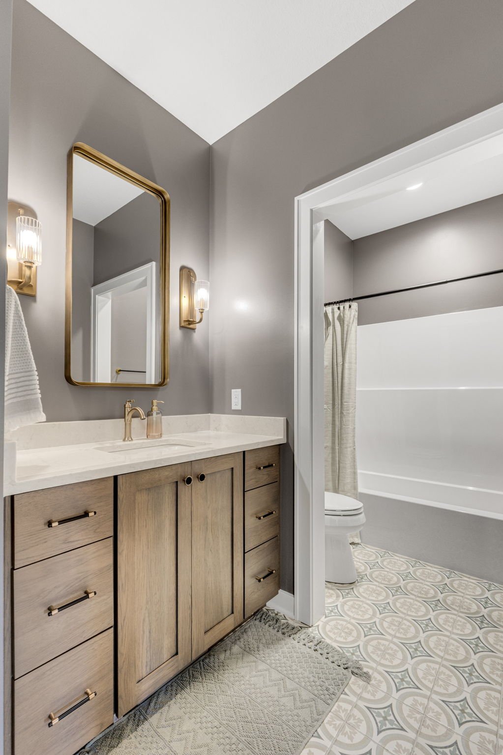 Modern bathroom with wooden vanity, beige countertop, gold fixtures, wall mirror, and decorative lighting, leading to a shower area with a white curtain and patterned floor tiles.