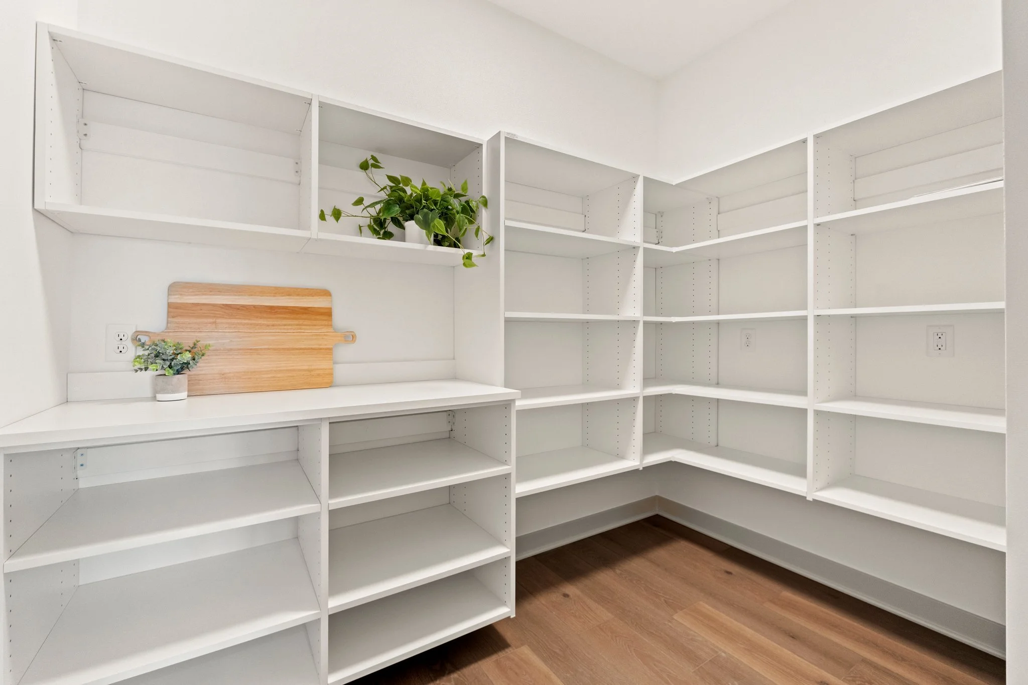Empty white corner pantry with built-in shelves, a small potted plant, and a cutting board, hardwood floor.