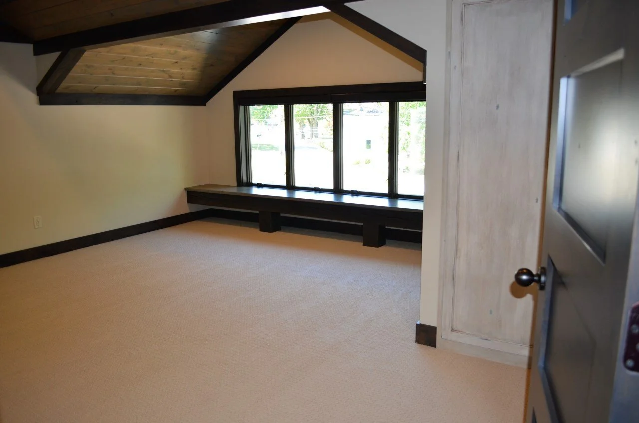 Empty room with a large window, beige carpet, and wood-paneled ceiling, partially viewed through an open door.