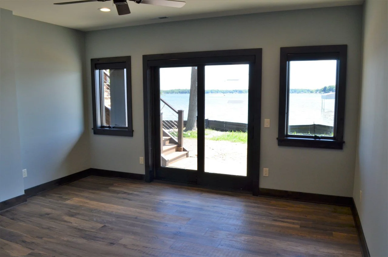 Empty room with wood flooring, gray walls, three windows, and a sliding glass door leading to an outdoor area with stairs, overlooking a body of water.