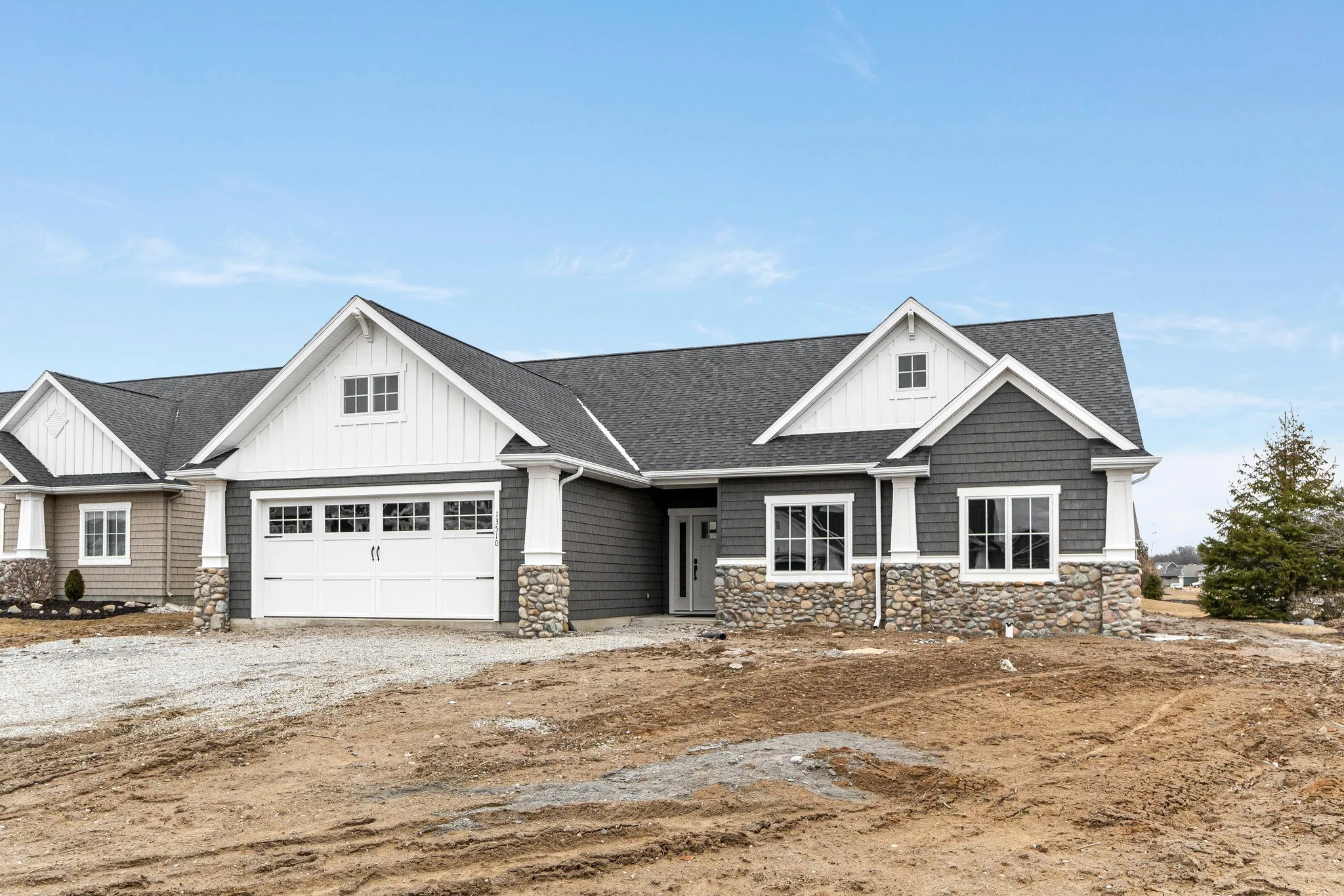 Newly built house with a gray exterior, white trim, stone accents, and a two-car garage with white doors, under a partly cloudy sky.