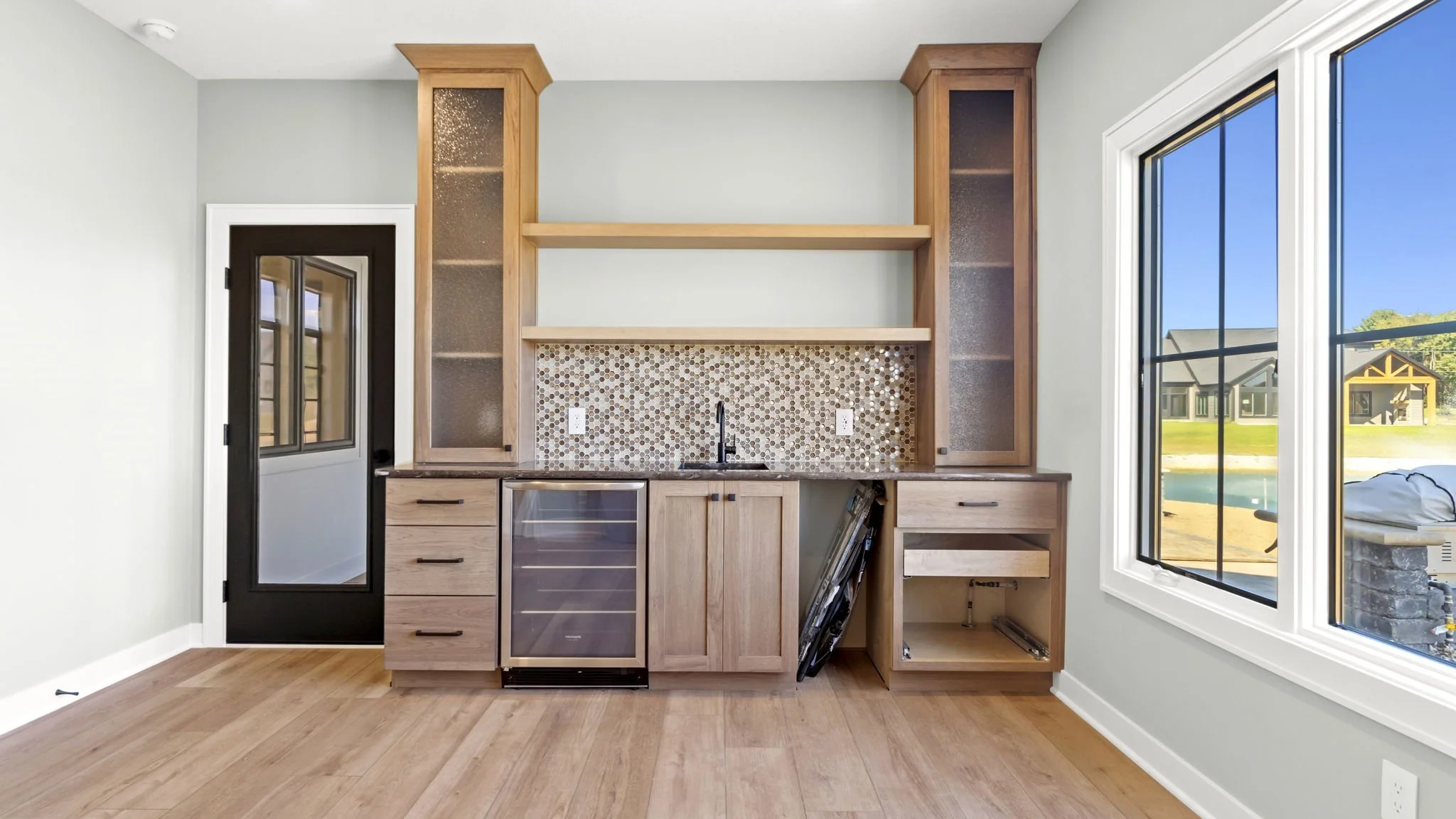 Kitchen with black door, large window, light wood cabinets, mosaic tile backsplash, and no appliances installed.