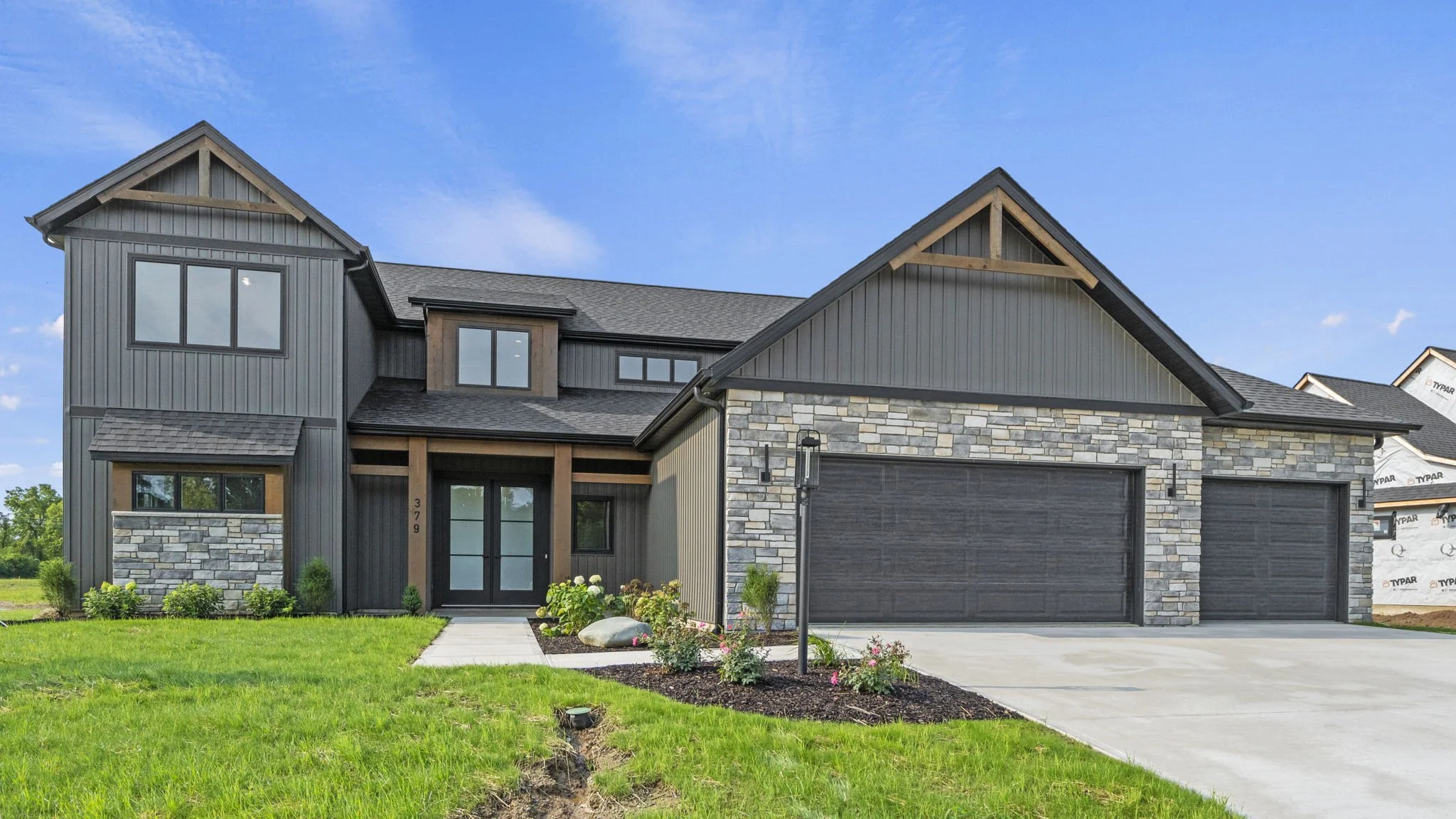 Modern two-story house with gray siding and stone accents, black garage doors, front lawn, and landscaped garden with flowers and shrubs, under a blue sky.