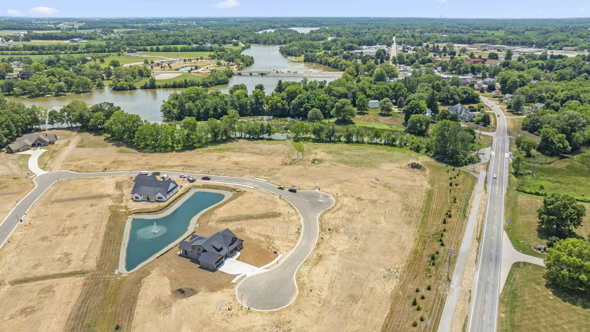 Aerial view of a residential area with two houses surrounded by a pond and a large body of water in the background, with trees, roads, and open land.