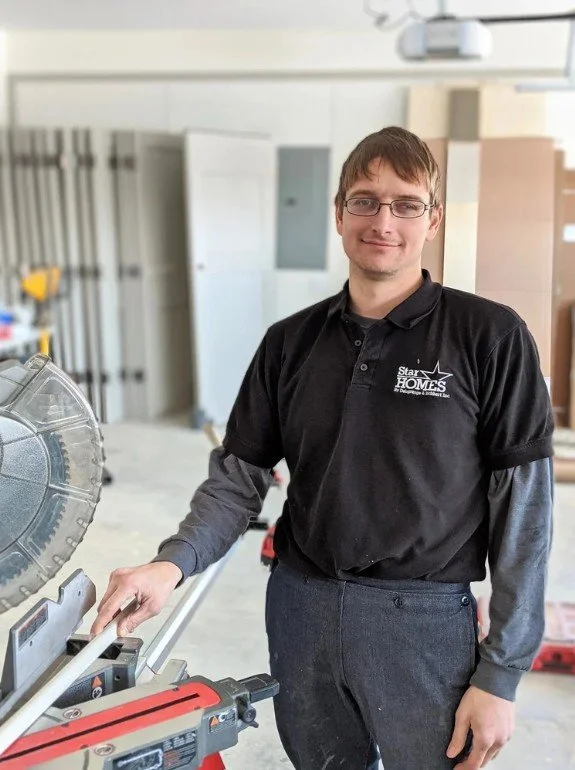 A man wearing glasses and a black shirt with a 'Start HOMES' logo stands in a workshop, holding a gray and red miter saw.