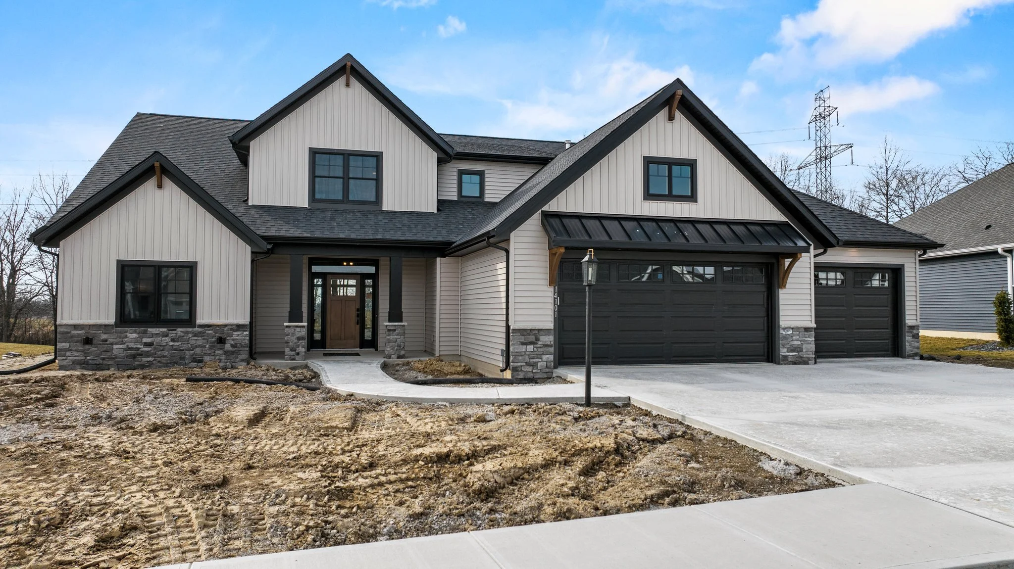 Newly built modern two-story house with a black garage door, stone and vinyl siding exterior, surrounded by a sidewalk and dirt yard, under a blue sky.