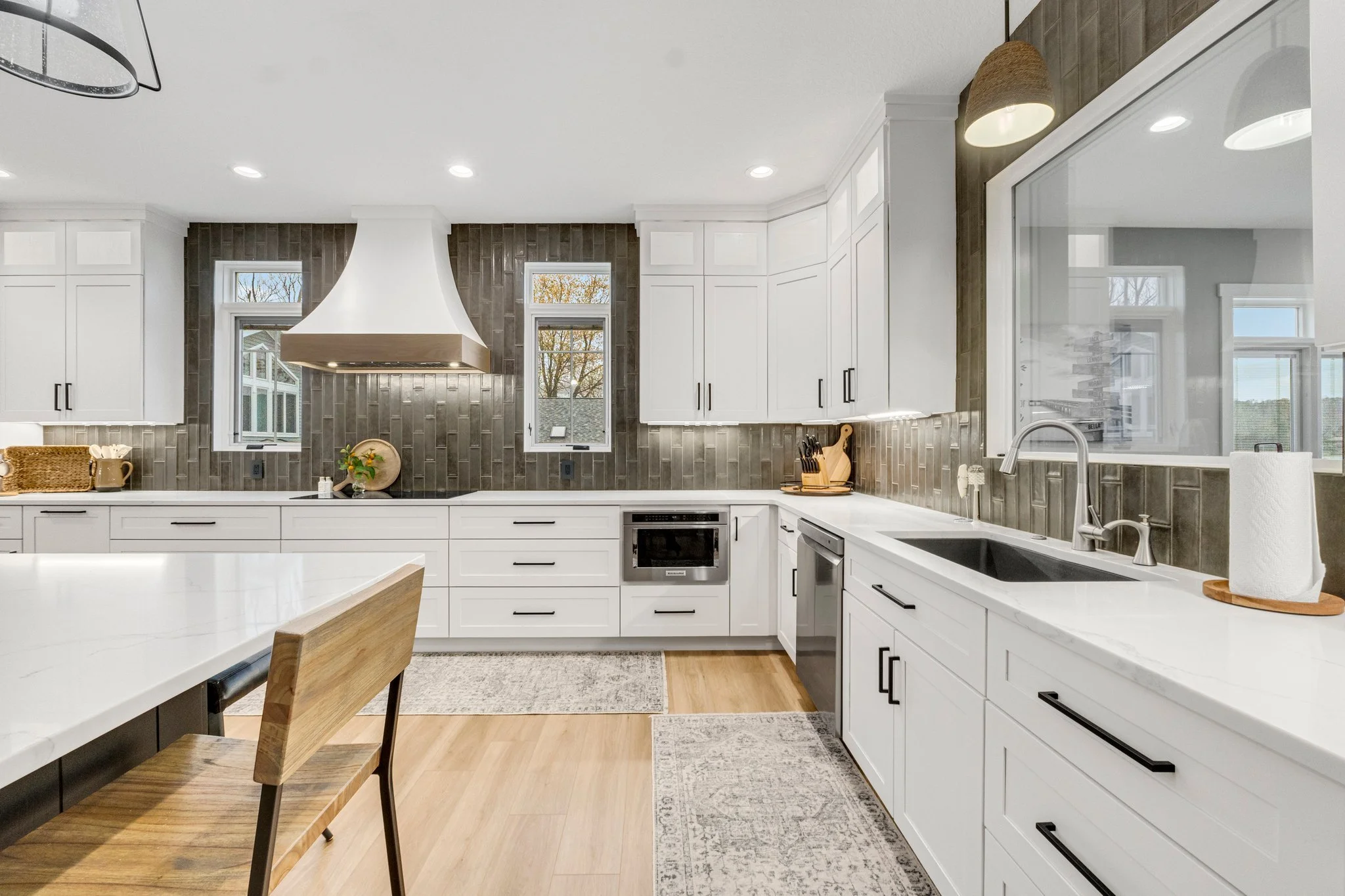 Modern kitchen with white cabinets, gray backsplash, and wooden accents, featuring a large window, island, and pantry.