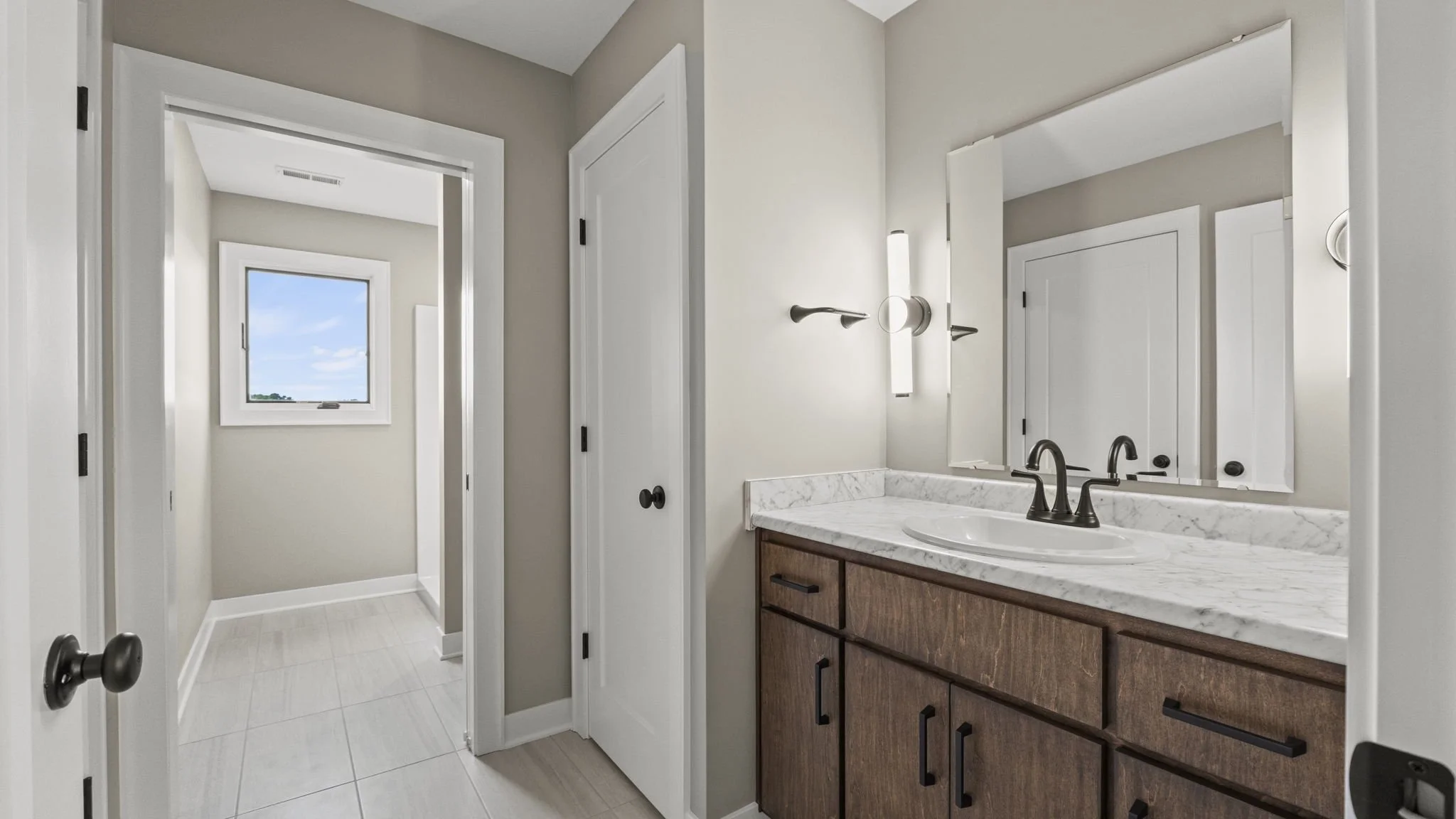 Bathroom with a wooden vanity with black handles, marble countertop, oval sink, wall-mounted faucet, large mirror, and modern light fixtures. Window with a view of blue sky and clouds visible in the background.