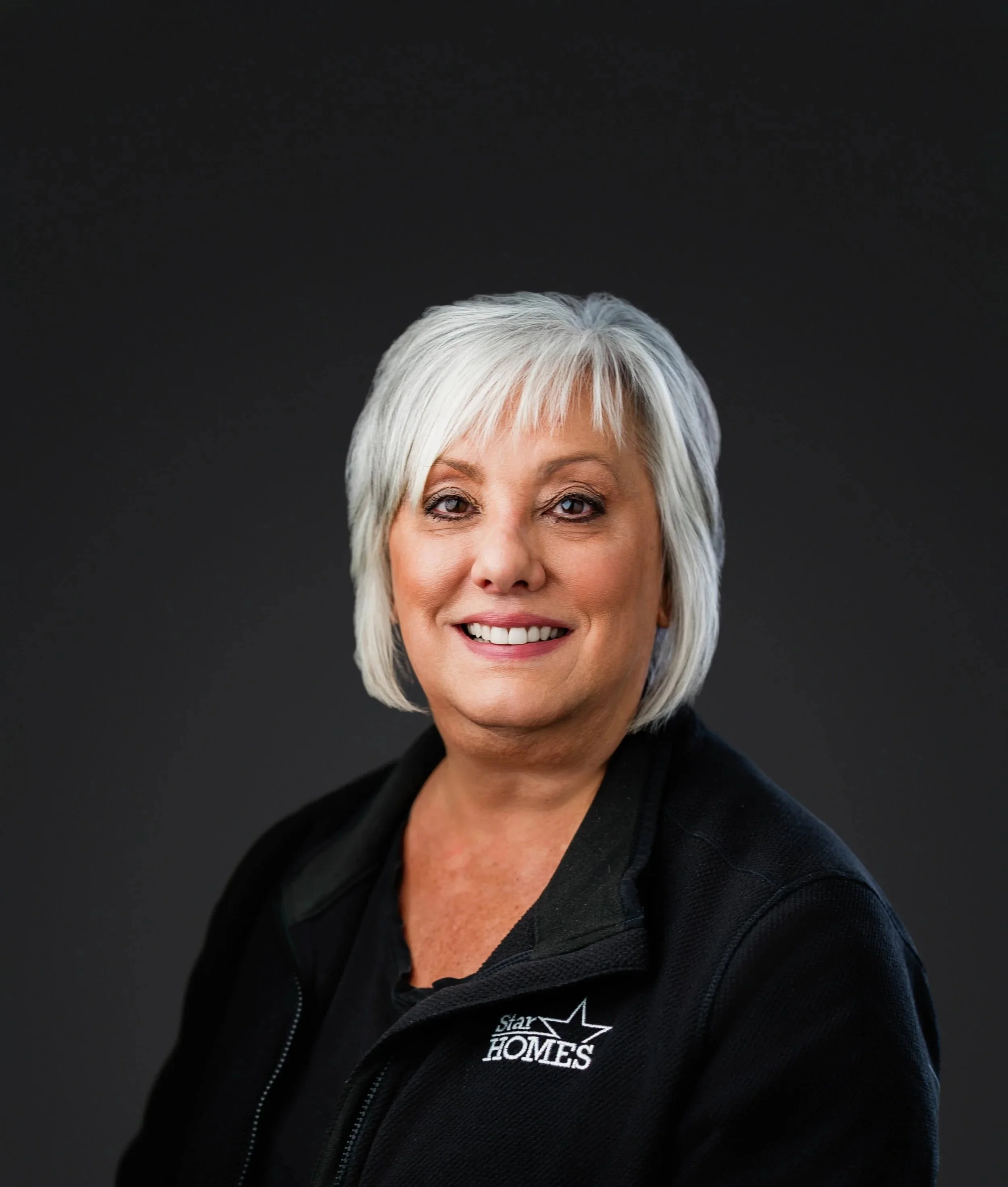 Close-up of a woman with short, silver hair, smiling and looking at the camera, wearing a black jacket with a logo that reads 'Star Homes' on it, against a black background.