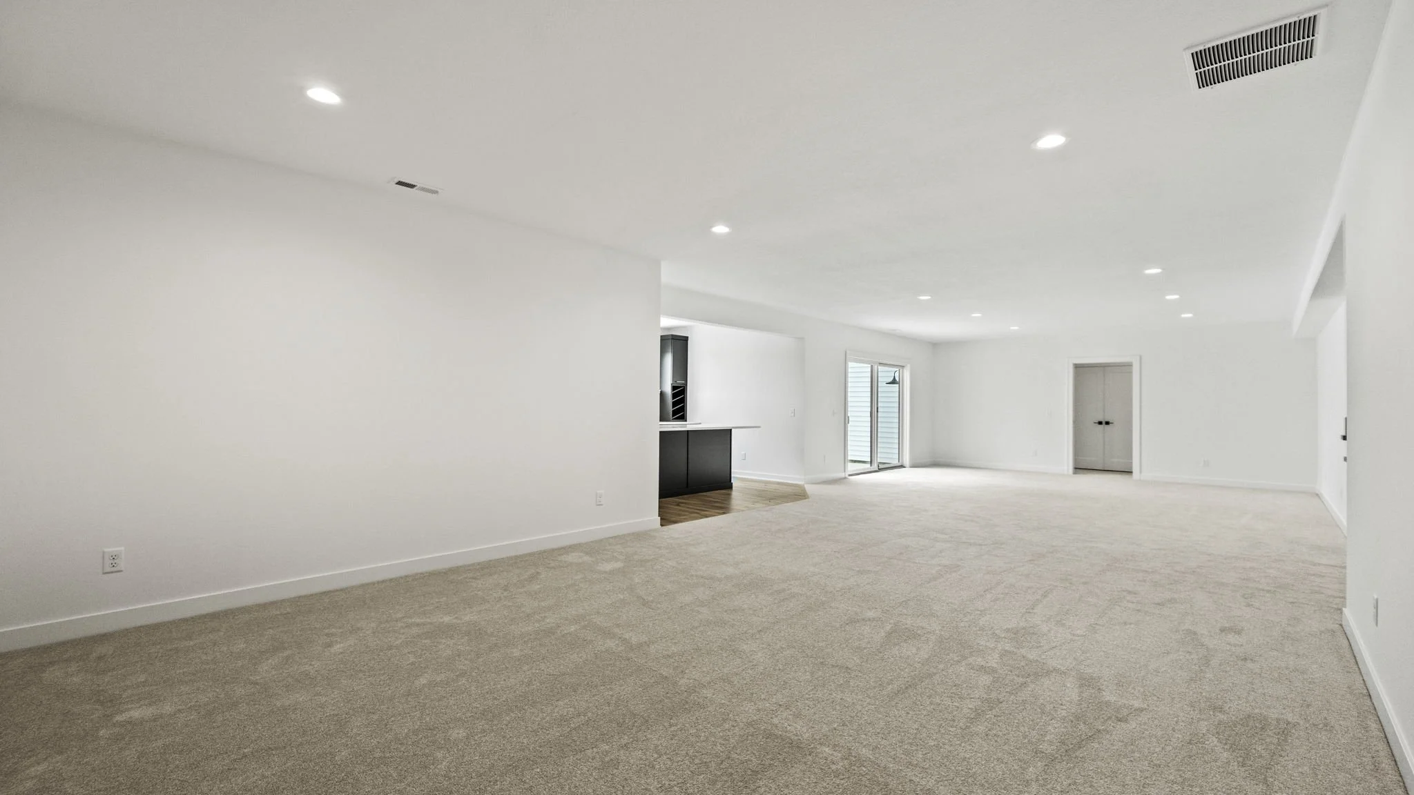 Spacious empty living room with beige carpet, white walls, and a white ceiling with recessed lighting, with sliding glass doors leading outside.