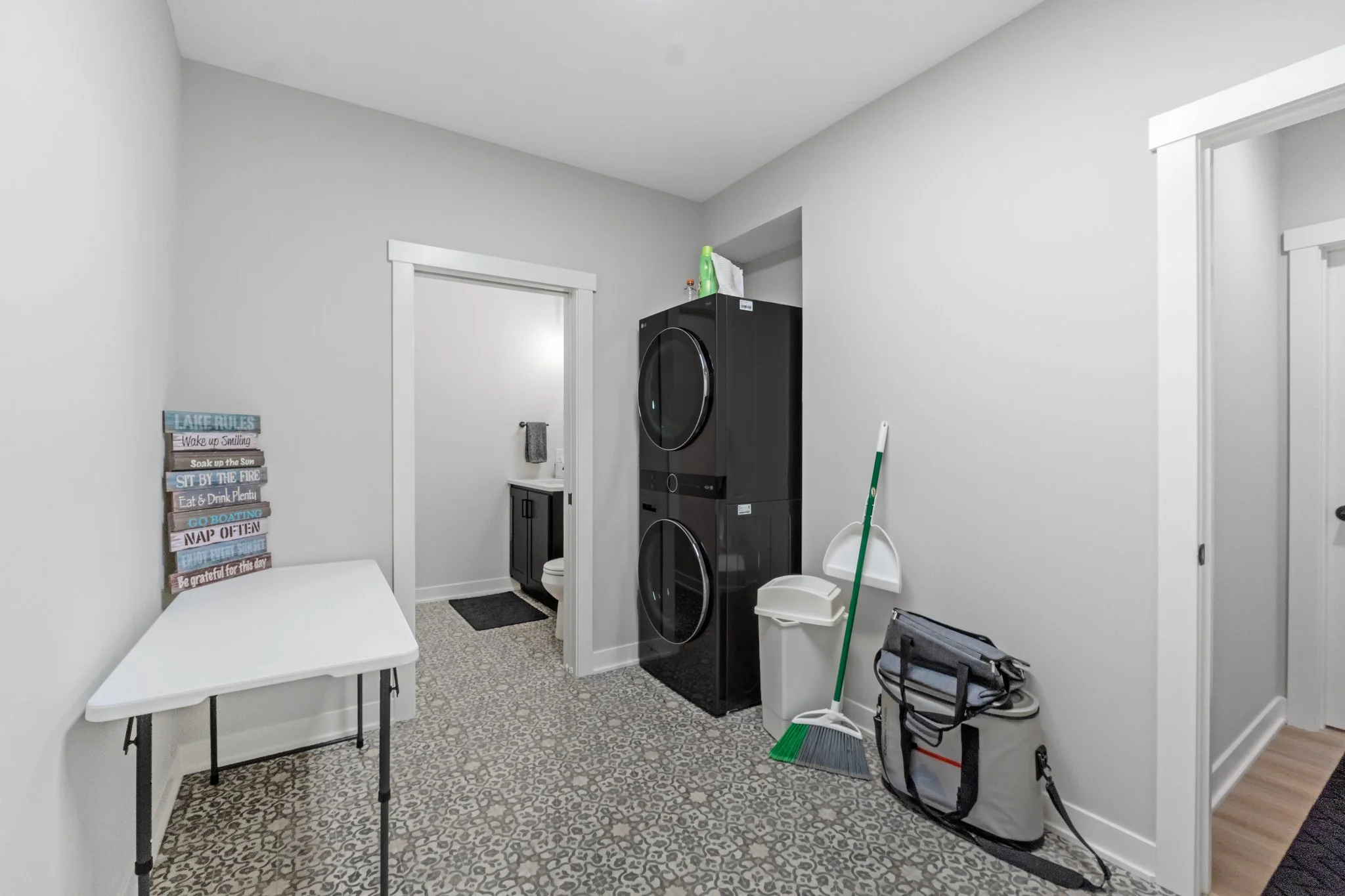 Laundry room with a stacked washer and dryer, a small table, cleaning supplies, a trash can, a broom, and a view of a bathroom with a vanity and toilet.
