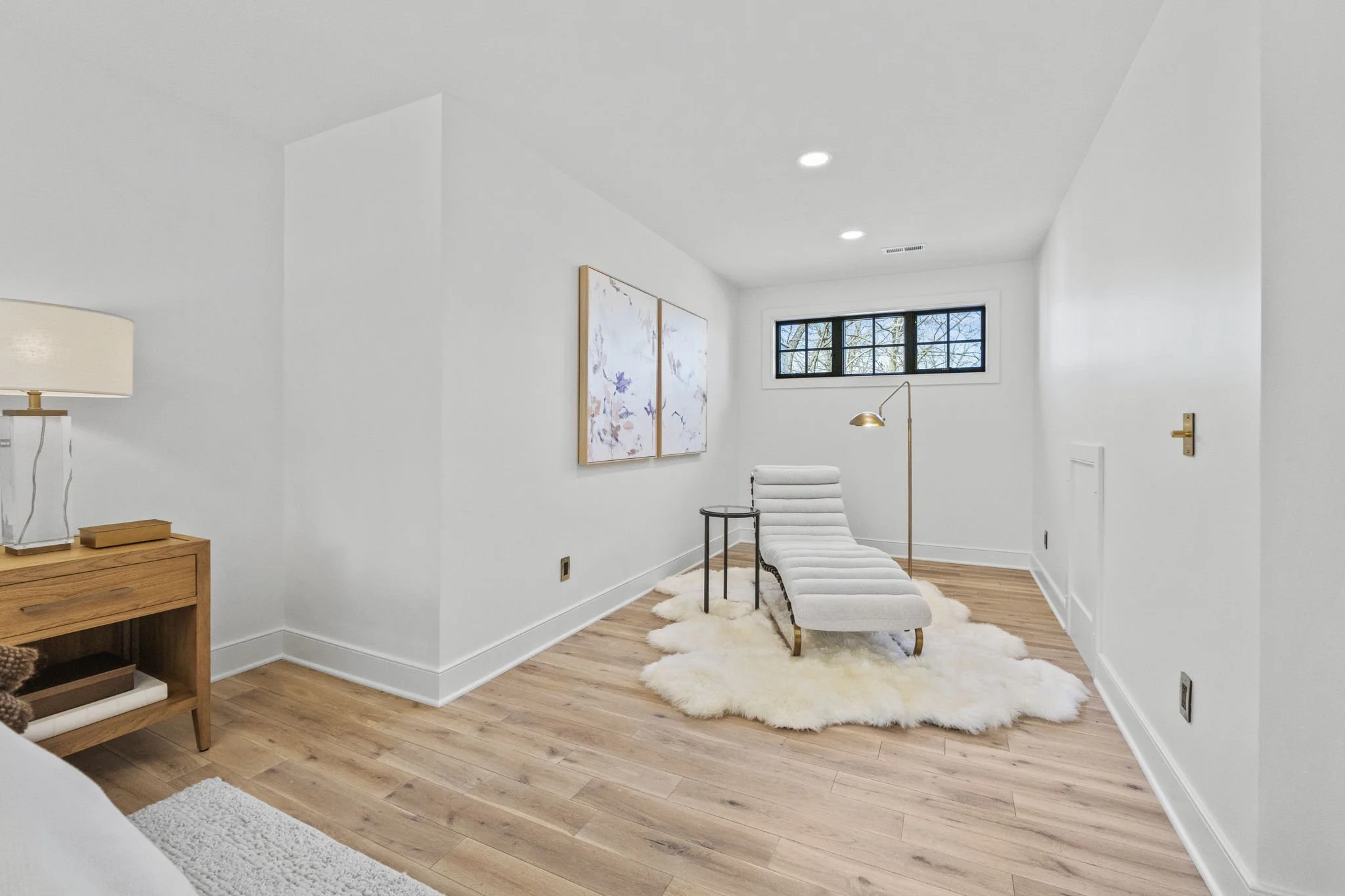 Minimalist white lounge chair on a fluffy cream rug in a bright, modern room with hardwood floors and white walls, featuring a window, wall art, and a gold floor lamp.