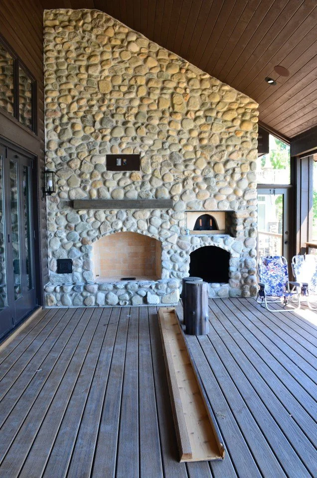 Interior view of a room with a large stone fireplace, wooden ceiling, and wooden deck. Sunlight is coming through large glass doors on the right.