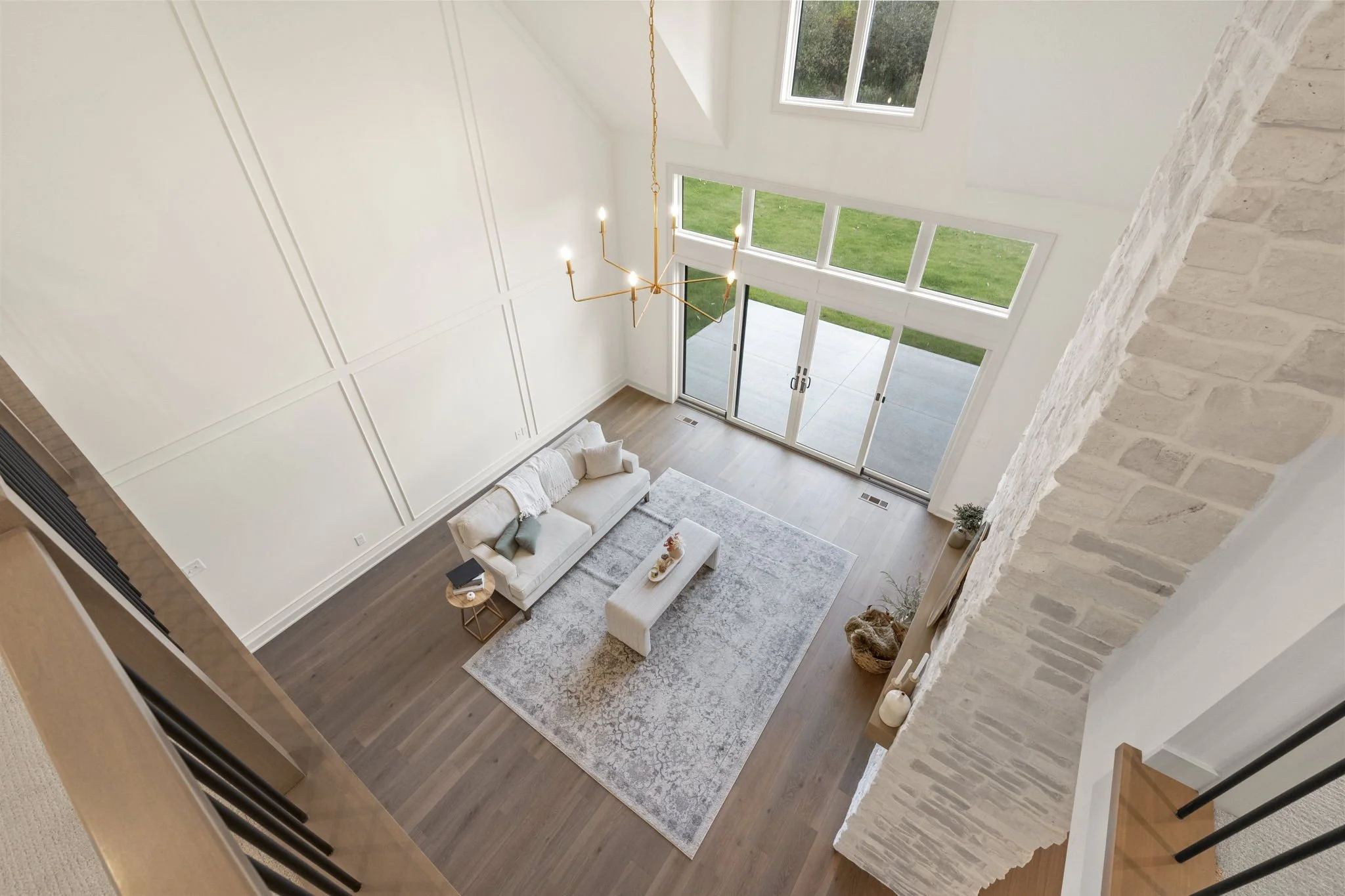 Living room with a high ceiling, large windows with a view of a green lawn, beige sofa with pillows, coffee table with decor, area rug, and a chandelier, taken from an upper vantage point.