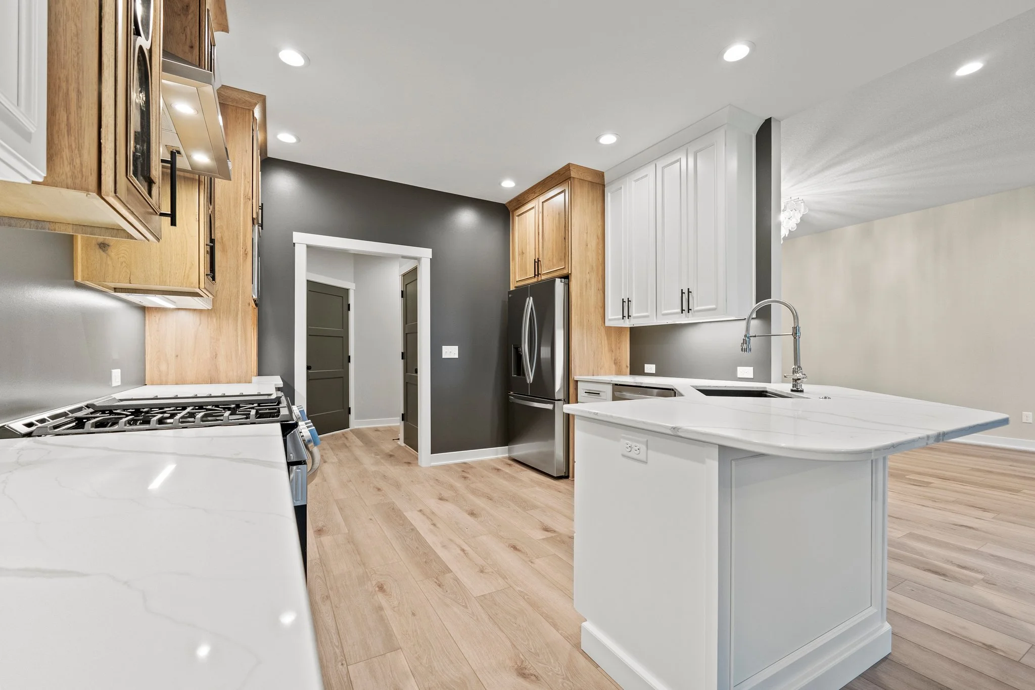 Modern kitchen with white and wood cabinets, stainless steel refrigerator, white countertops, and a kitchen island with a sink. Light wood flooring, gray and white walls, and recessed lighting.
