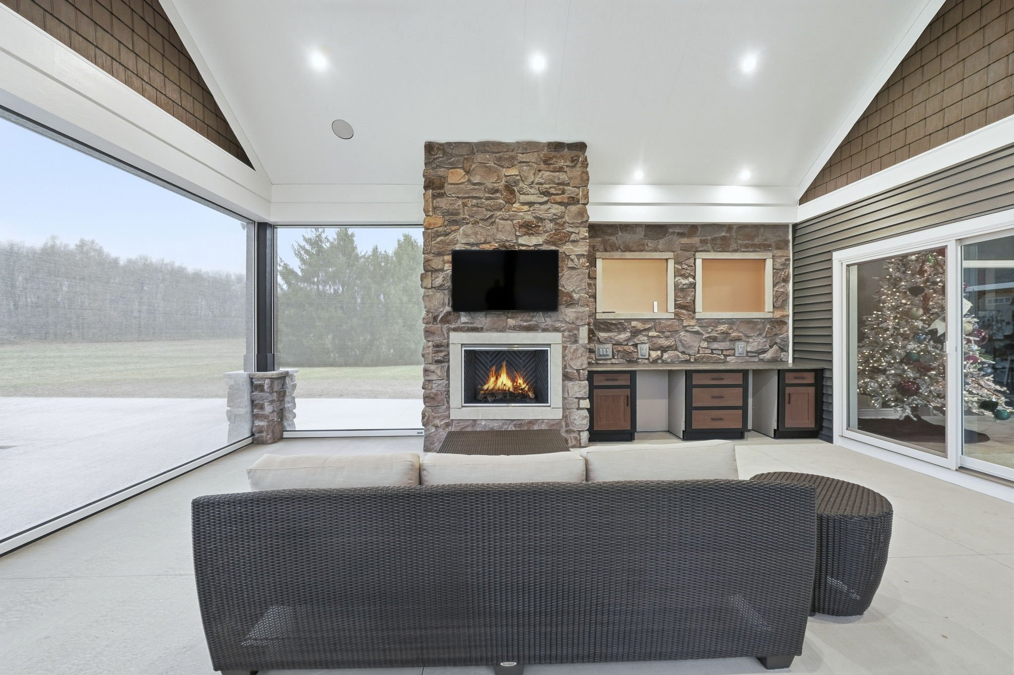 Modern living room with stone fireplace, wall-mounted TV, beige sofa, wicker chair, sliding glass doors, and Christmas tree visible through the window.