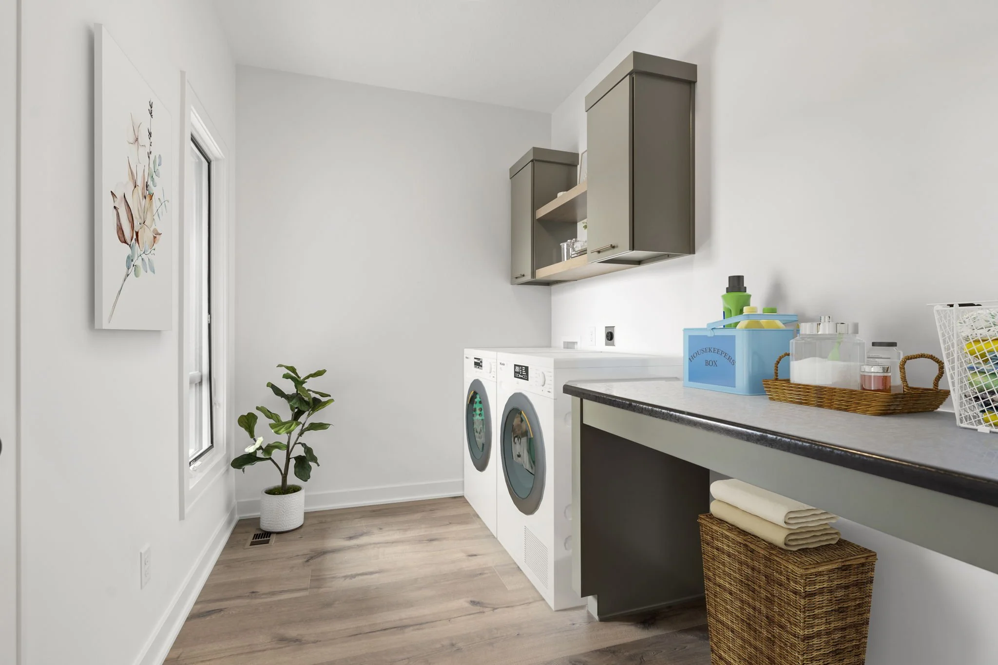 Laundry room with white walls, wooden floor, a potted plant near a window, gray cabinets and countertop, washing machine and dryer, basket of laundry, and cleaning supplies.