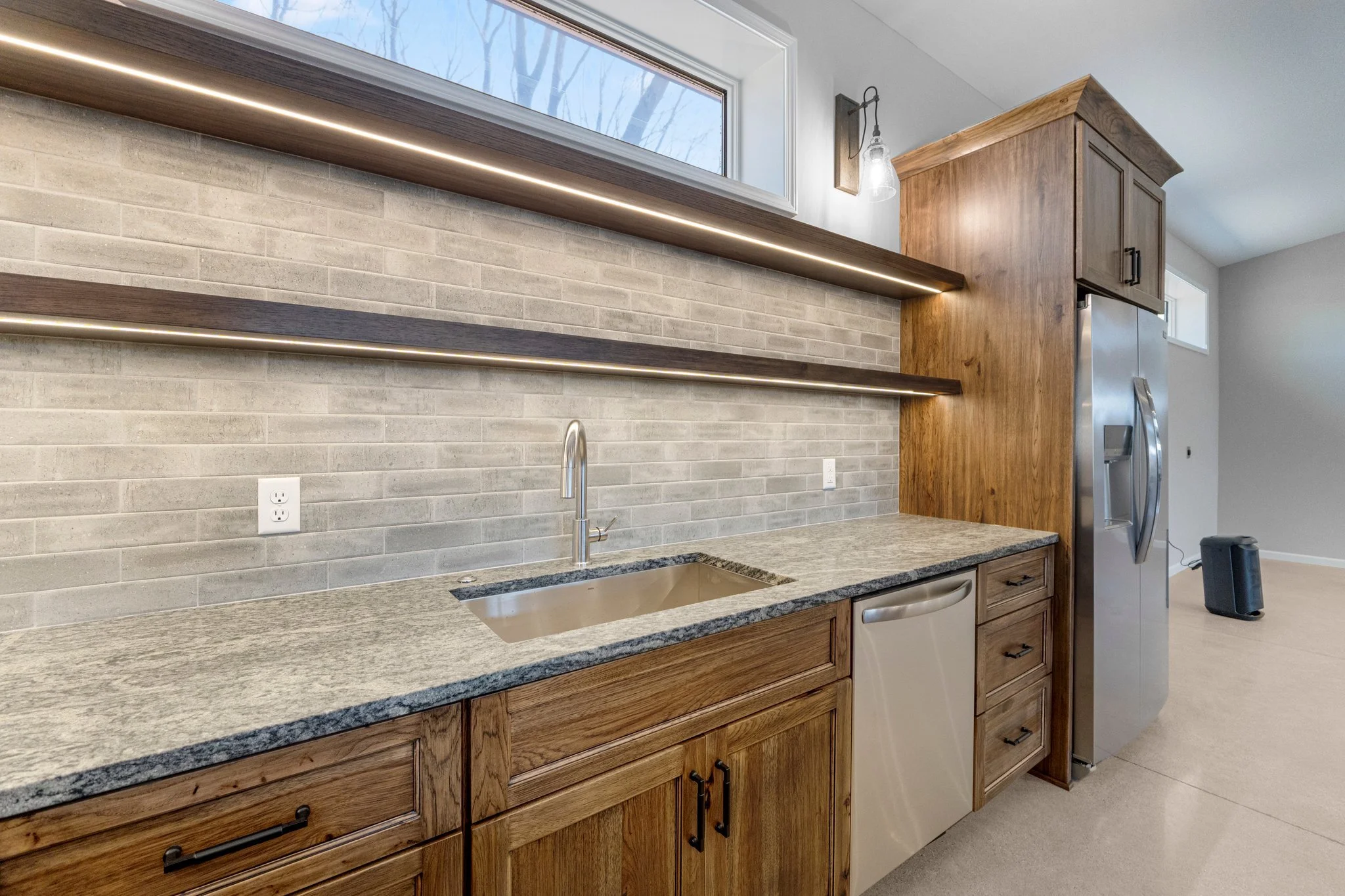 Kitchen with wooden cabinets, granite countertop, stainless steel sink, brick backsplash, open floating shelves, and a refrigerator.