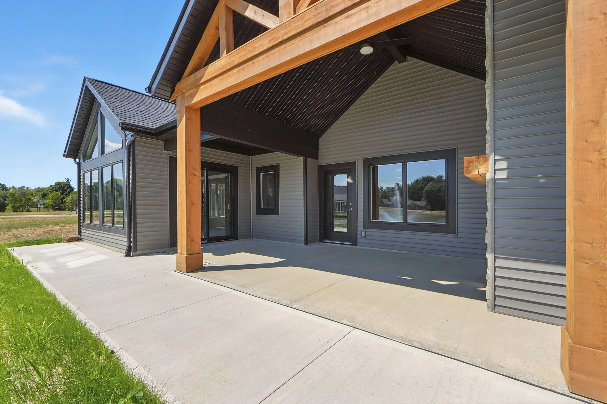 Exterior view of a house with a covered patio, grey siding, black window frames, and a concrete floor. Wooden beams support the patio roof.