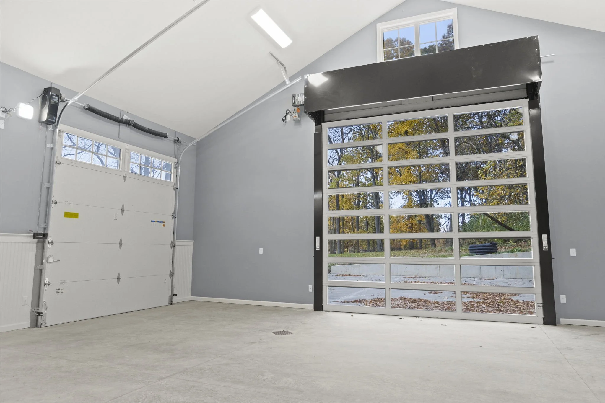 Empty garage with two large doors, one white and one transparent, and gray walls, with trees visible outside.