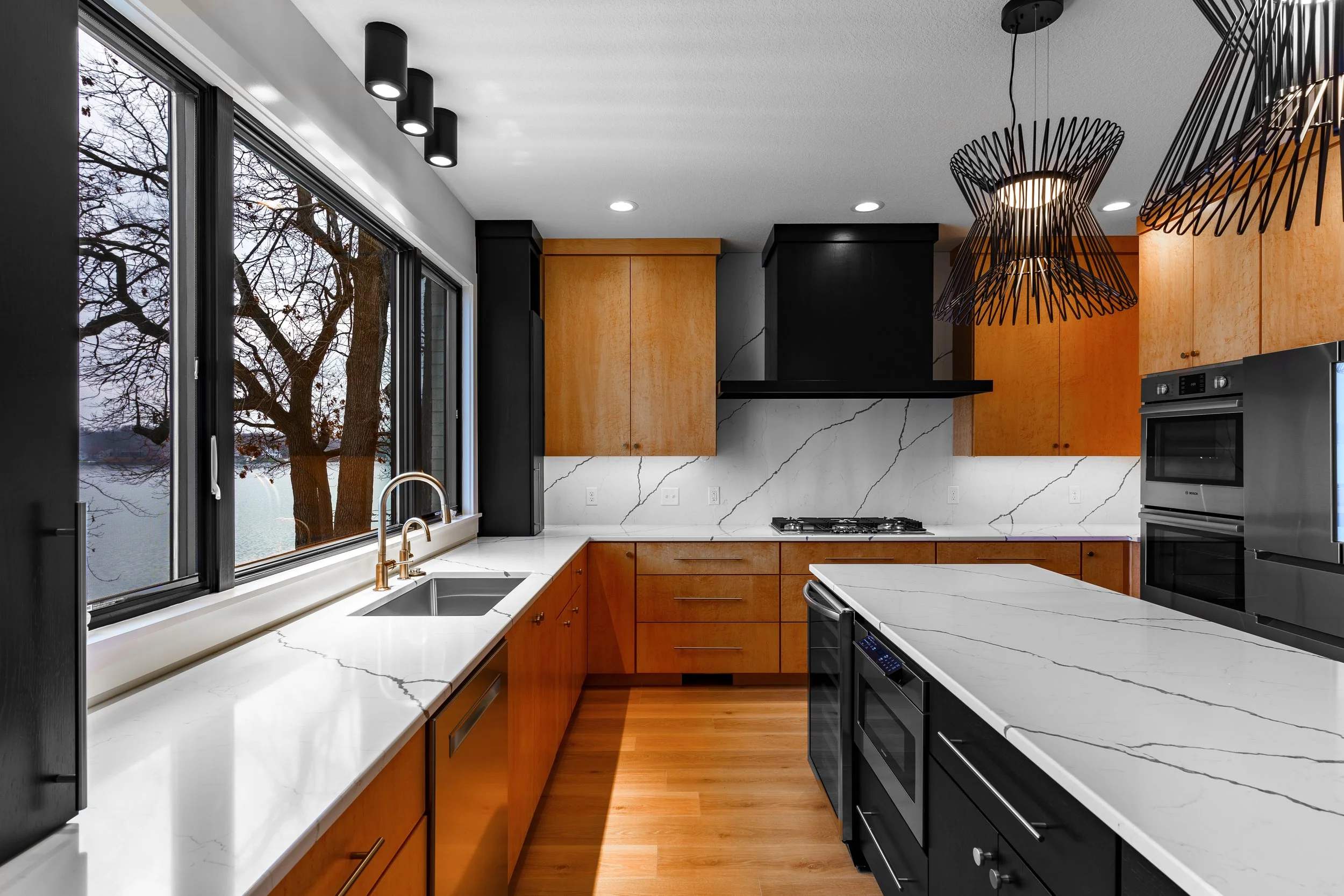 Modern kitchen with large window, white marble countertops with black veining, wooden cabinets, black and stainless steel appliances, pendant lights, and a view of trees outdoors.