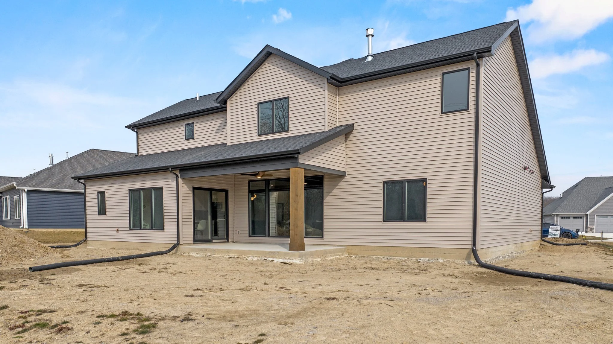 A newly built, beige-colored, multi-story house with large windows and a covered back patio, situated on a plot of land with bare soil, with neighboring houses visible in the background under a blue sky.