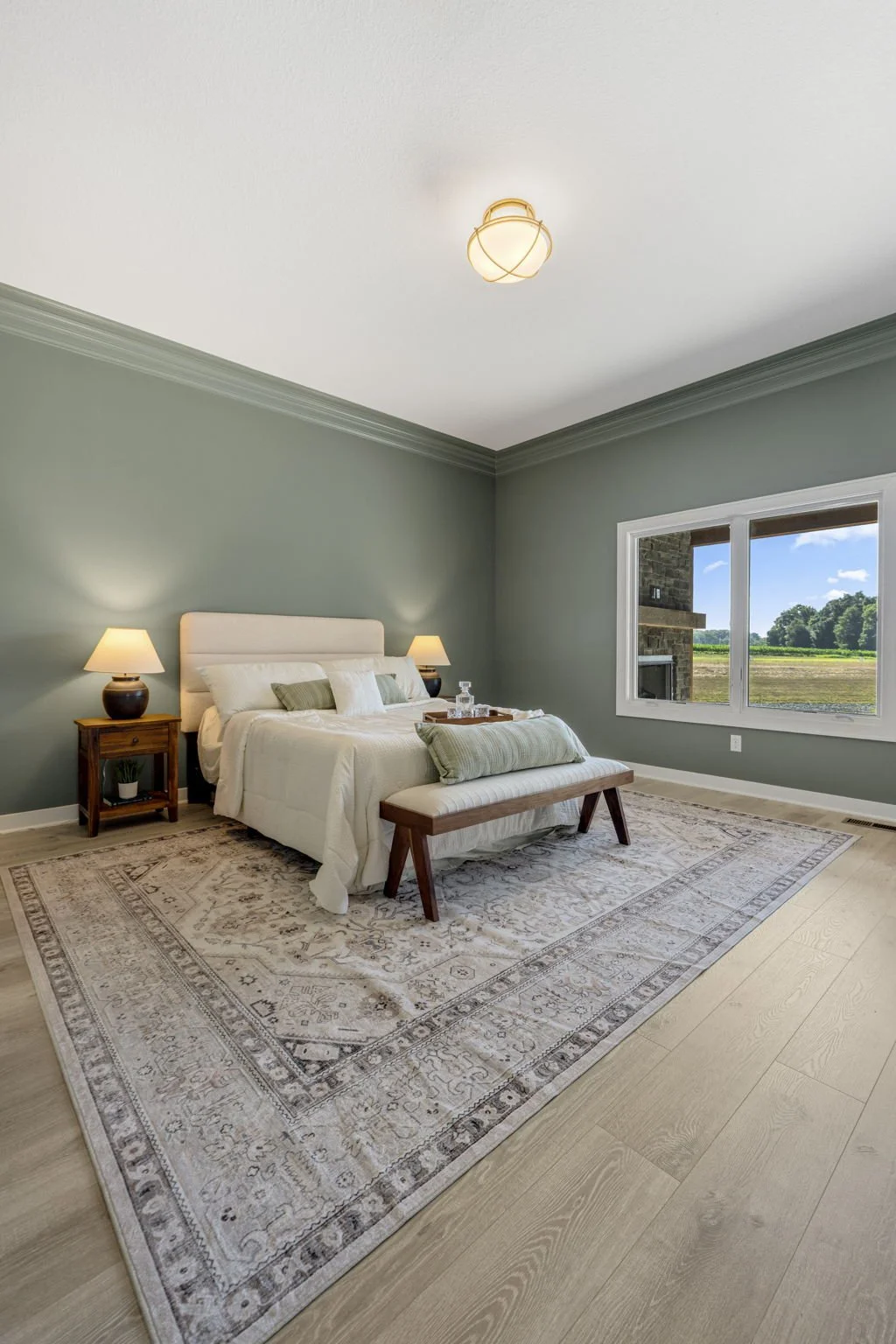 A bedroom with green walls, a white bed with pillows, two nightstands with lamps, a bench at the foot of the bed, a large window showing an outdoor view, and a patterned rug on light wood flooring.
