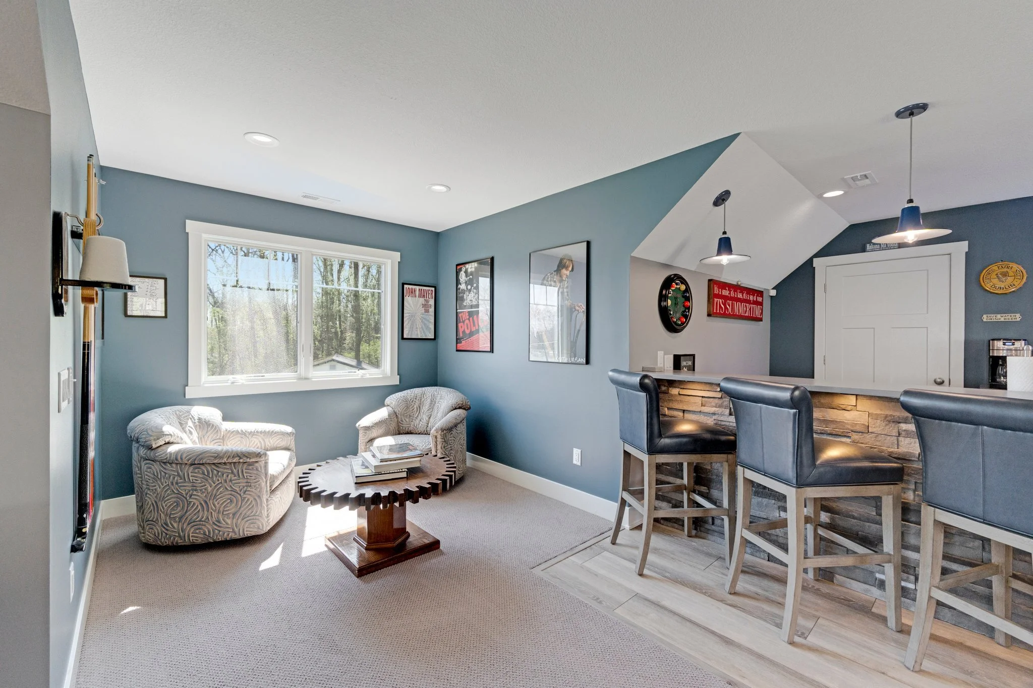 A cozy living room with blue walls, a large window, two patterned armchairs, a wooden coffee table, and a bar area with three bar stools, pendant lights, and framed posters on the wall.
