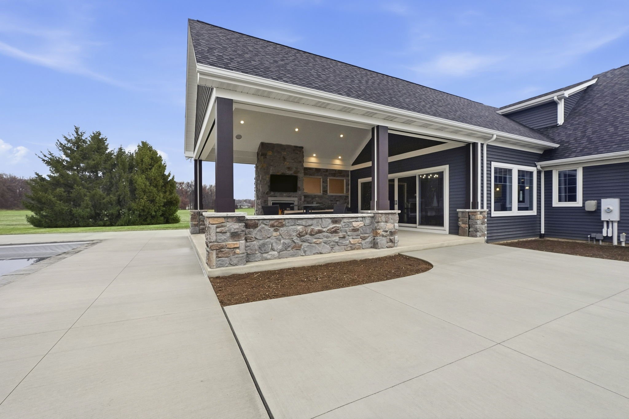 Backyard patio with concrete flooring, a covered outdoor living area with stone and dark wood accents, a mounted flat-screen TV, and a built-in fireplace, with lawn and trees in the background.