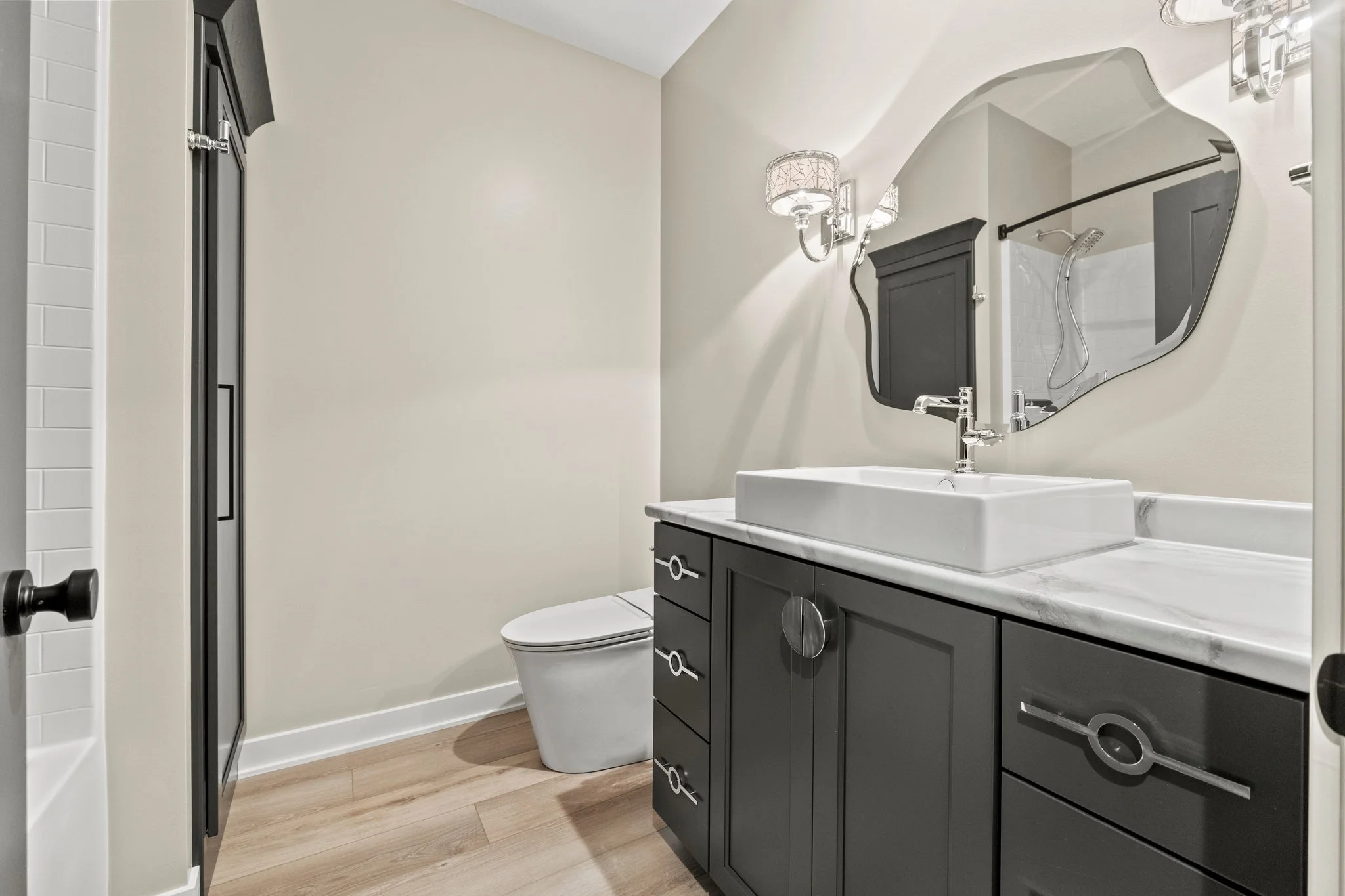 Modern bathroom with a white toilet, black vanity with silver handles, white marble countertop, square white vessel sink, large mirror, black wall-mounted cabinet, wall-mounted silver light fixture, and a walk-in shower with a glass door and black fr