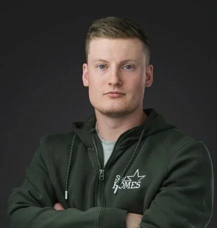 Young man wearing a dark green hoodie with crossed arms, facing the camera against a dark background.