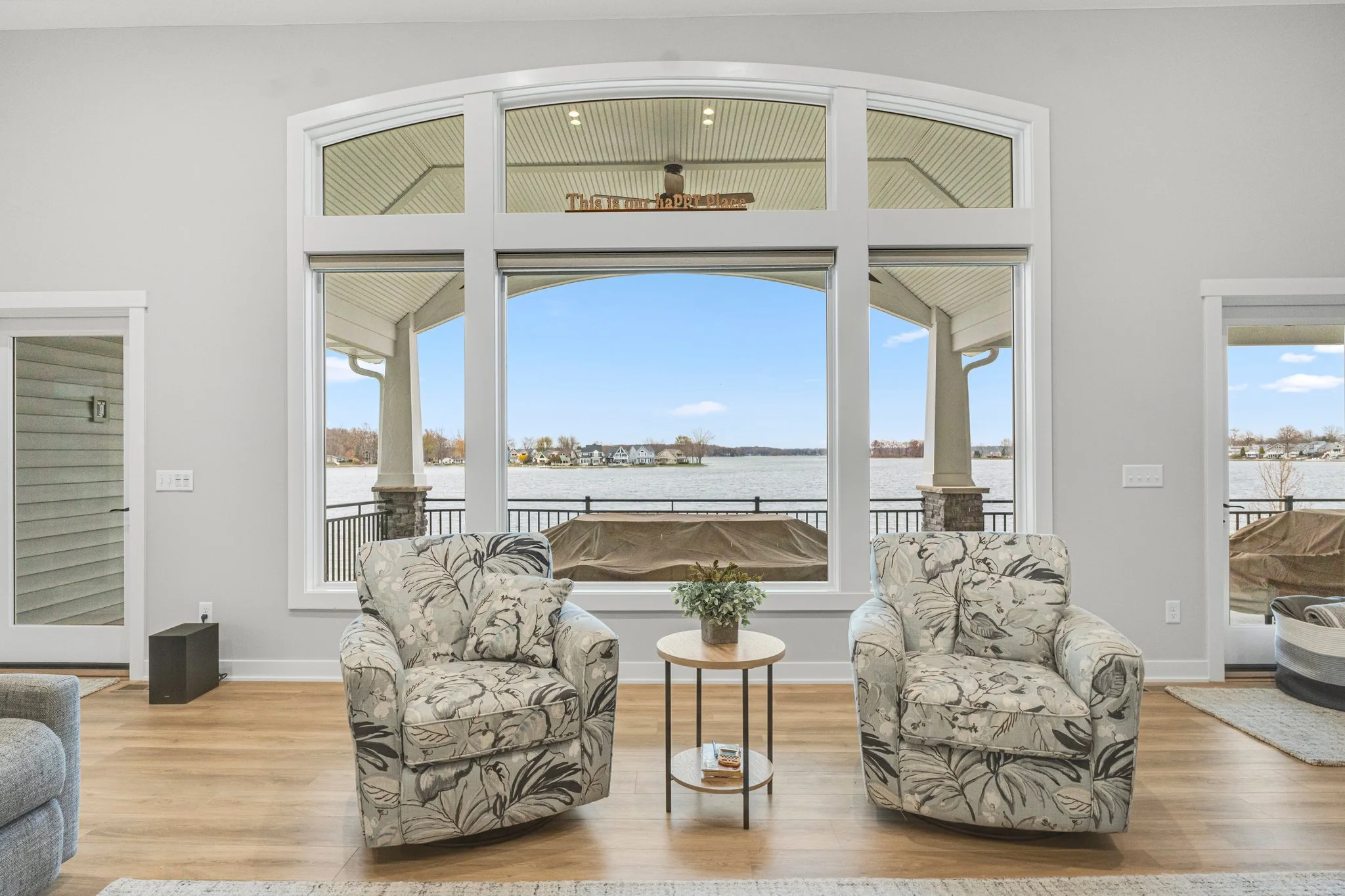Living room with large window showing lake view, two floral armchairs, small round table with plants, hardwood floor.