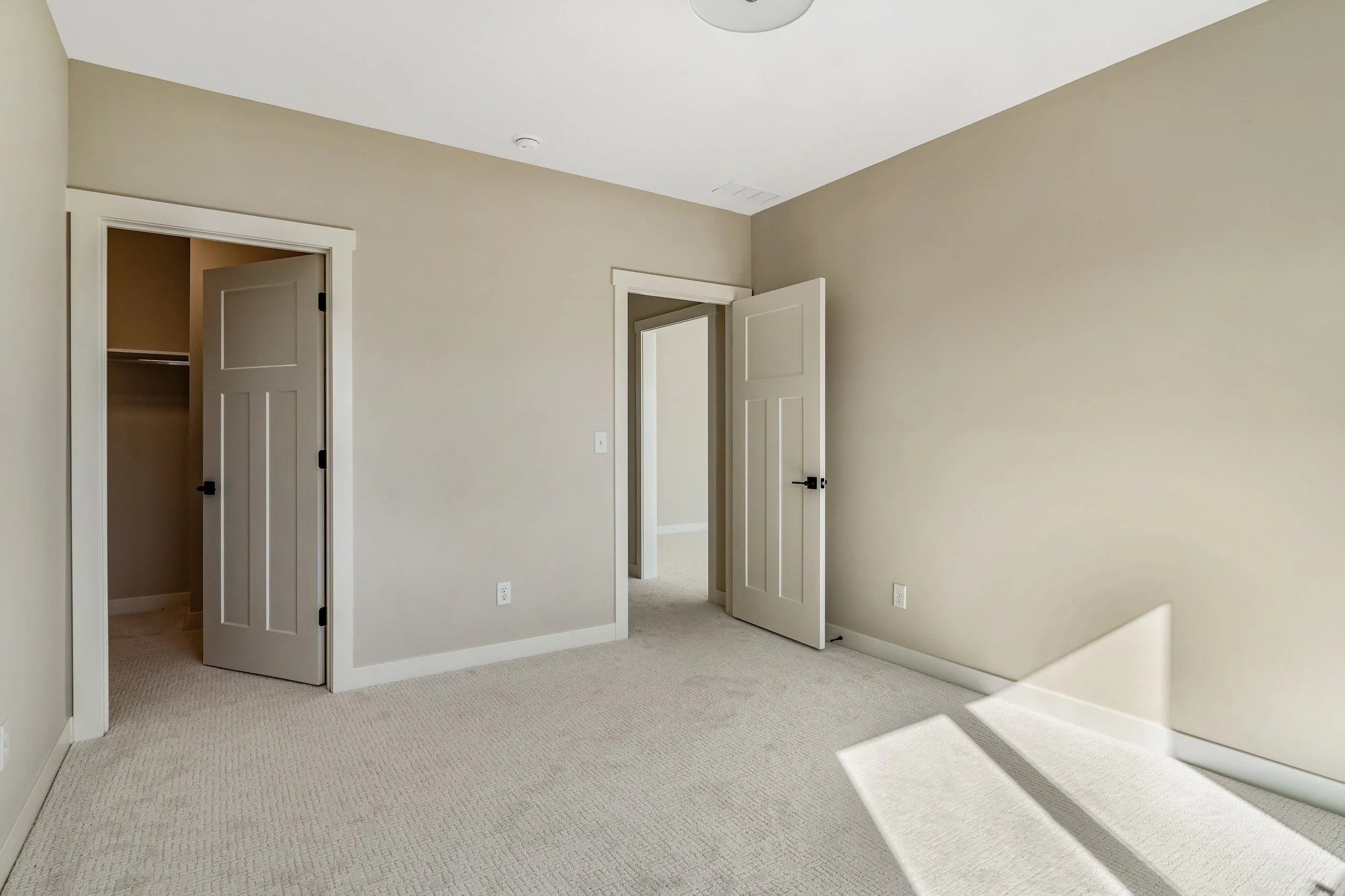 Empty bedroom with beige walls, beige carpet, and white trim. Two doors, one leading to a walk-in closet, and one open door, with sunlight streaming in creating a shadow on the floor.